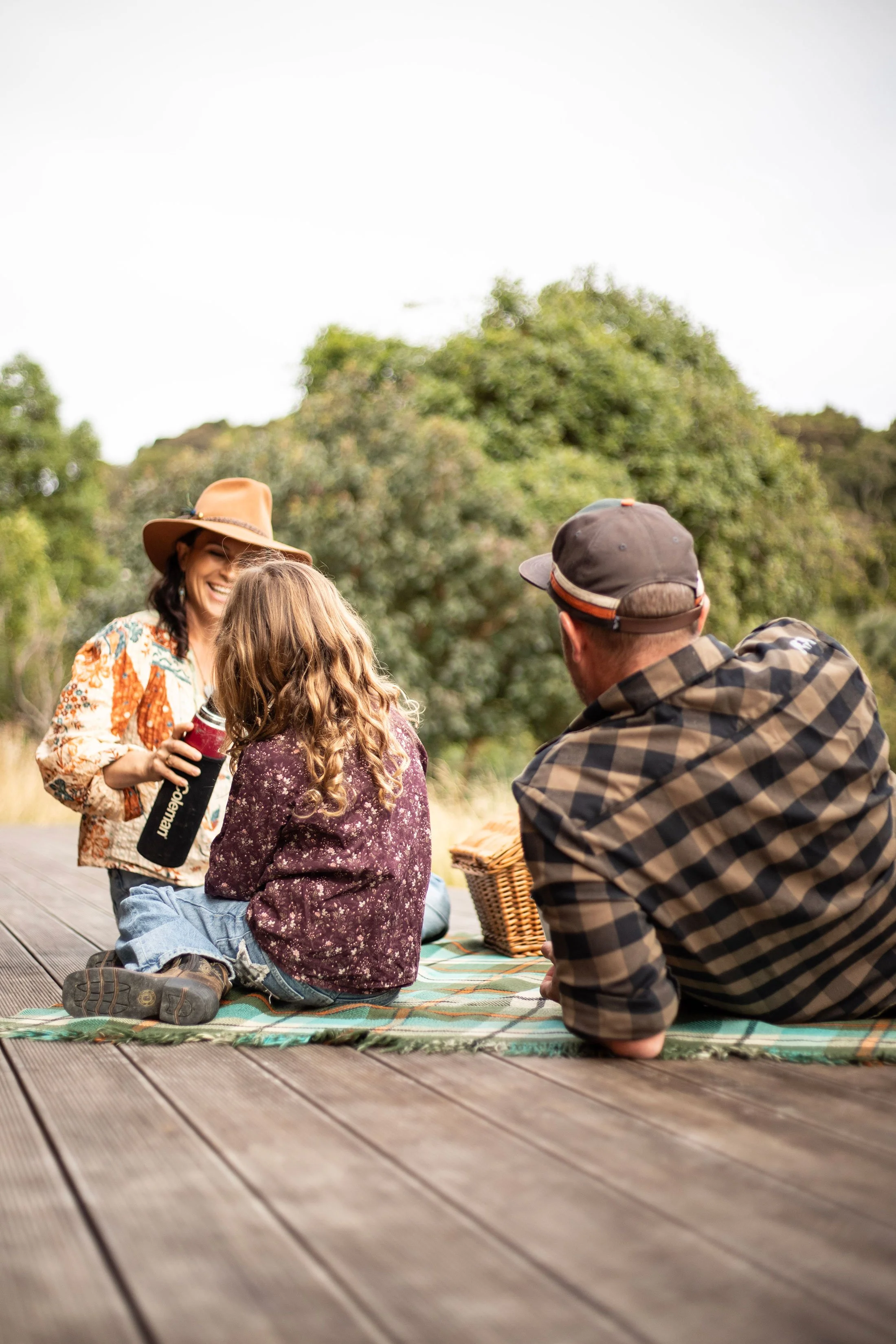 A woman wearing a wide-brimmed hat and colorful blouse smiling and talking to children and a man sitting on a picnic blanket outdoors, with trees in the background.