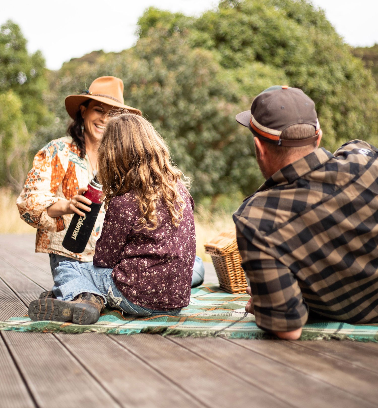 A woman wearing a wide-brimmed hat talking with a man and a girl sitting on a blanket outdoors, with trees in the background. They are enjoying a conversation and smiling.
