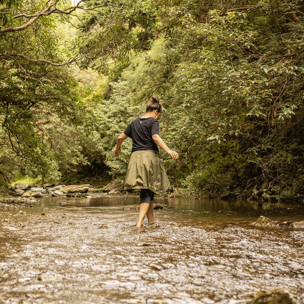 A woman walking through a shallow creek surrounded by dense green trees in a forest.