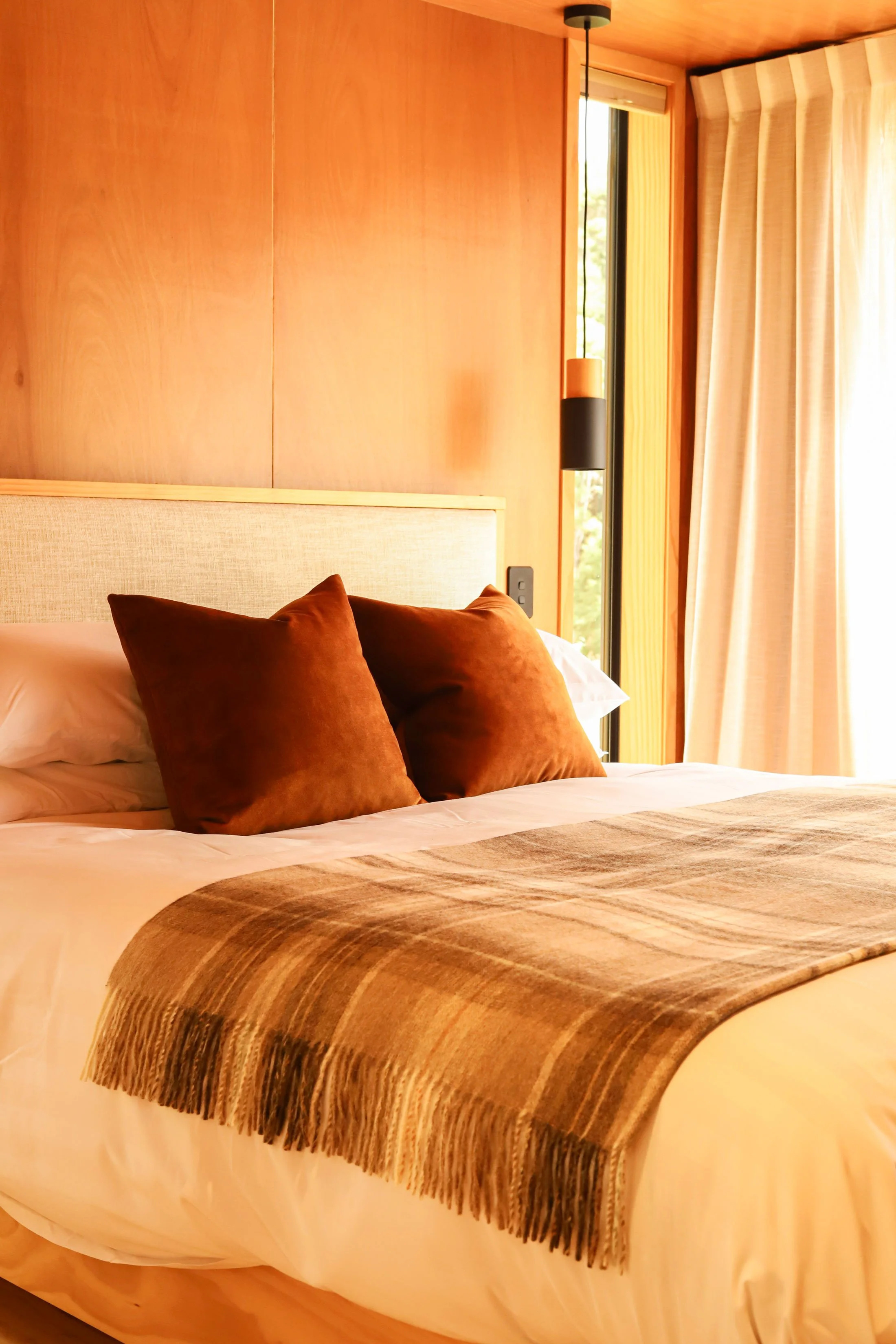 A neatly made bed with rust-colored pillows and a fringed brown, striped blanket on top. The headboard is beige, and there are wooden walls and a window with cream-colored curtains in the background, along with a hanging light fixture.