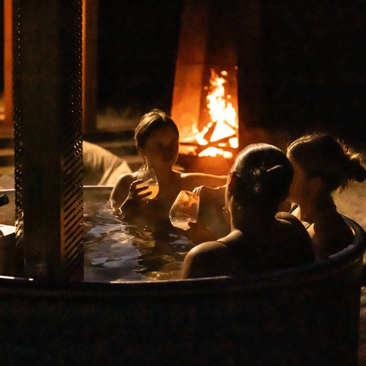 Three women relaxing in a hot tub at night, with a fire pit in the background.