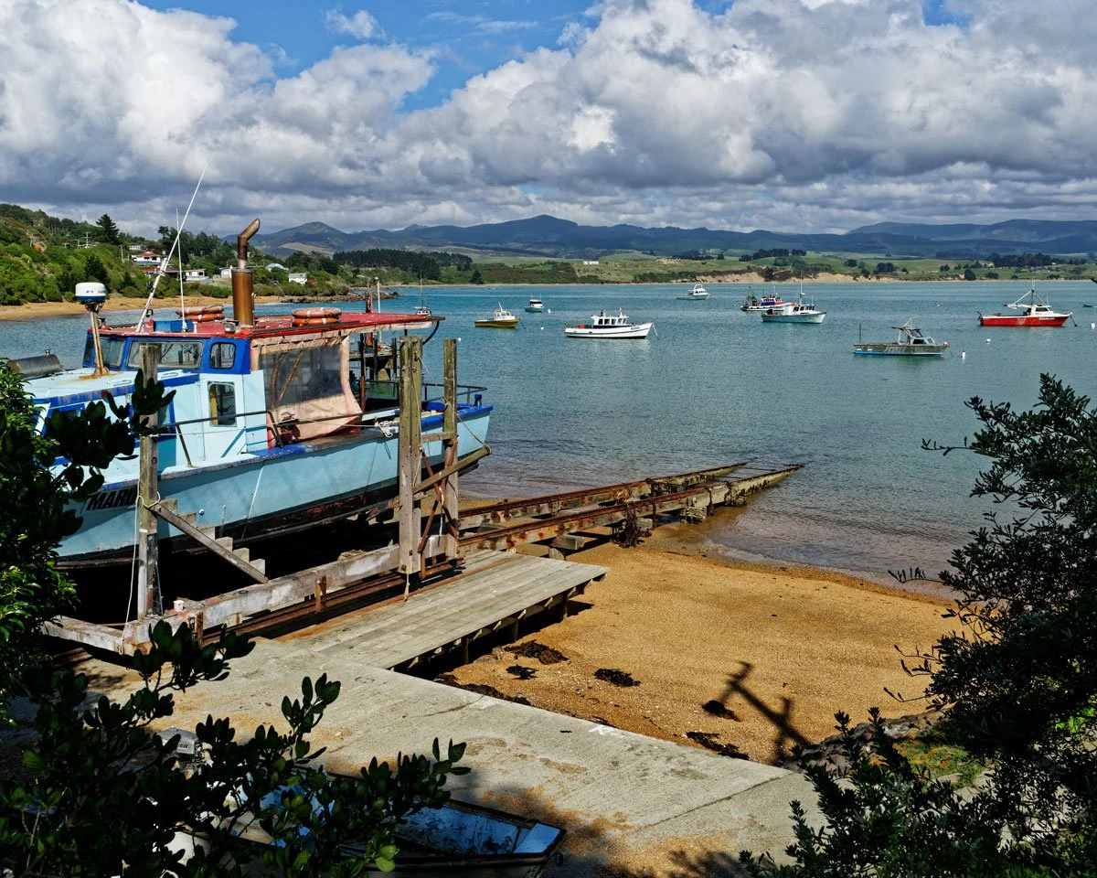 A boat on a wooden cradle at a lakeside, with a sandy beach and calm water holding several other boats, green hills, and mountains under a partly cloudy sky.