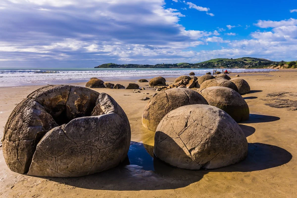 Large round boulders on a sandy beach with waves and green hills in the background under a partly cloudy sky.