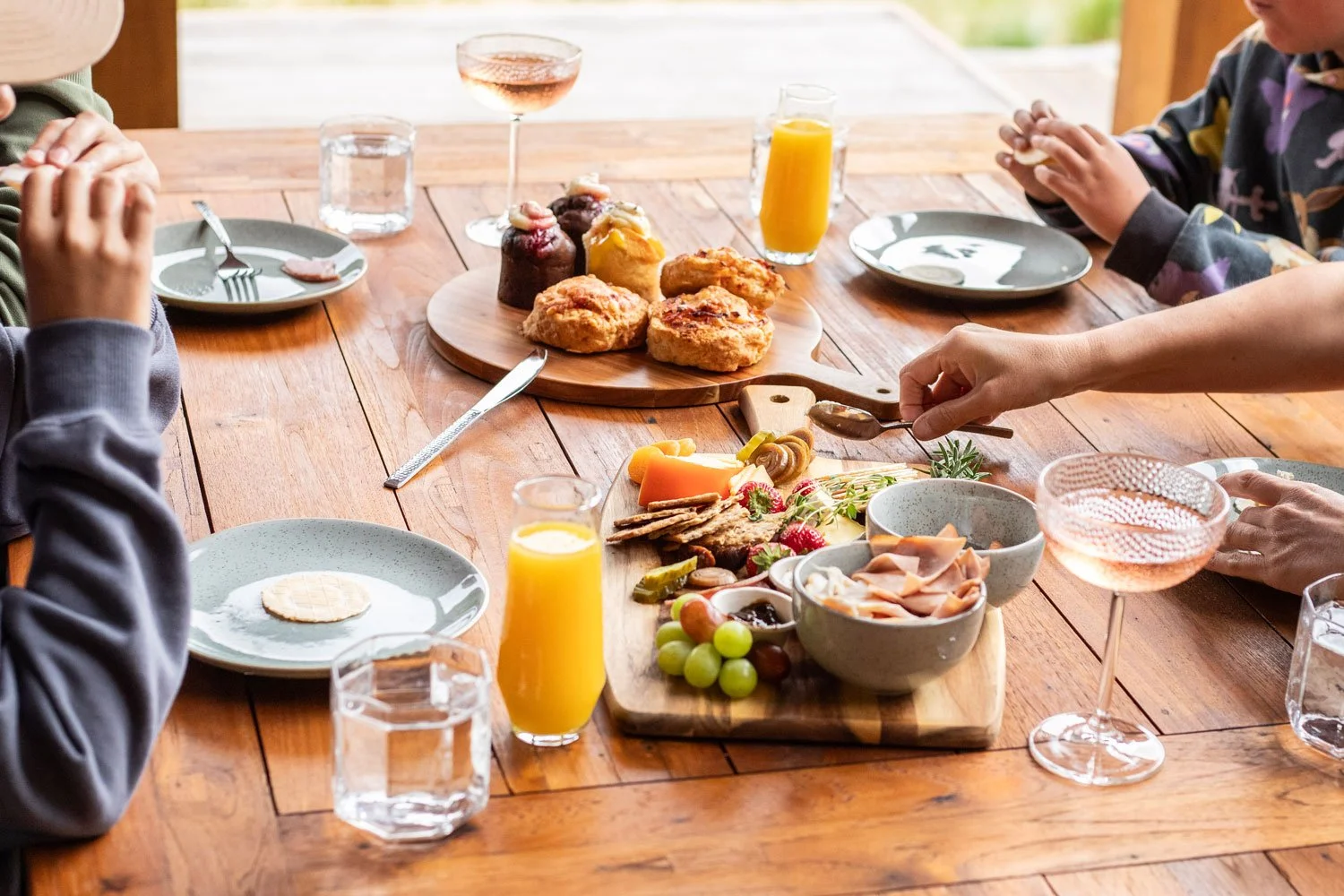 A family breakfast spread on a wooden table with assorted pastries, fruits, cheese, cold cuts, and glasses of orange juice and rosé wine.
