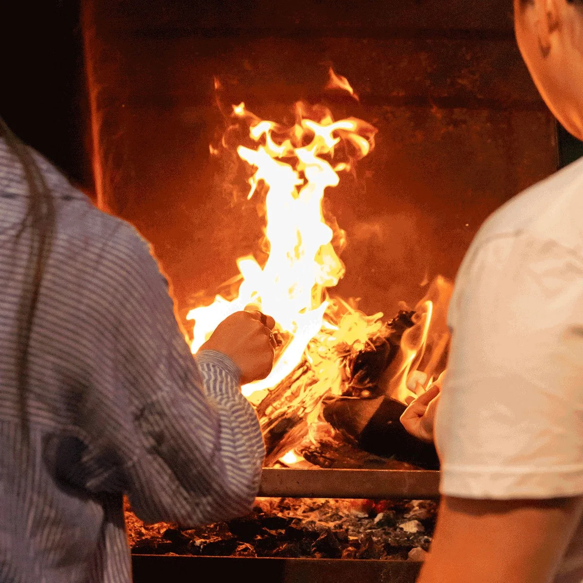 Two people sitting by a fireplace, tending to the fire with their hands.