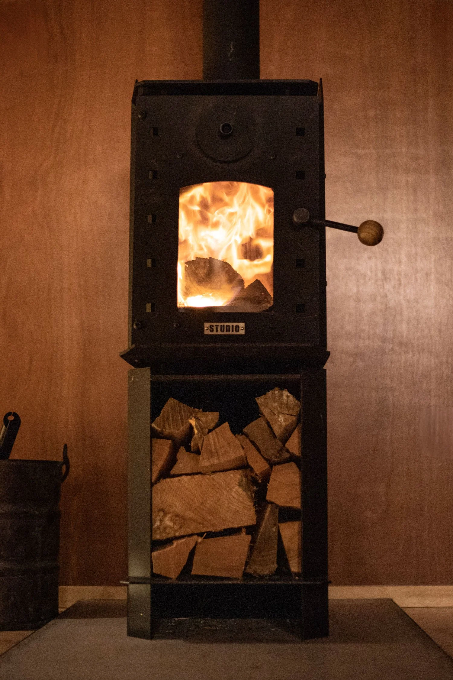 A modern indoor wood-burning stove with a fire inside, positioned above a storage area filled with chopped firewood, against a textured wood-paneled wall.