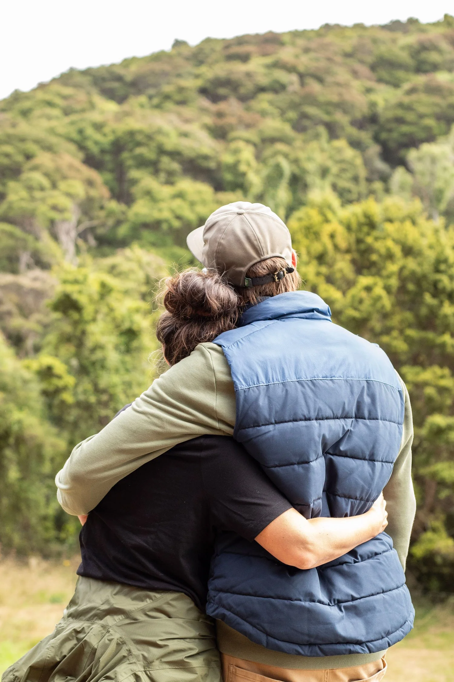 A couple embracing outdoors in front of a green, wooded hillside.