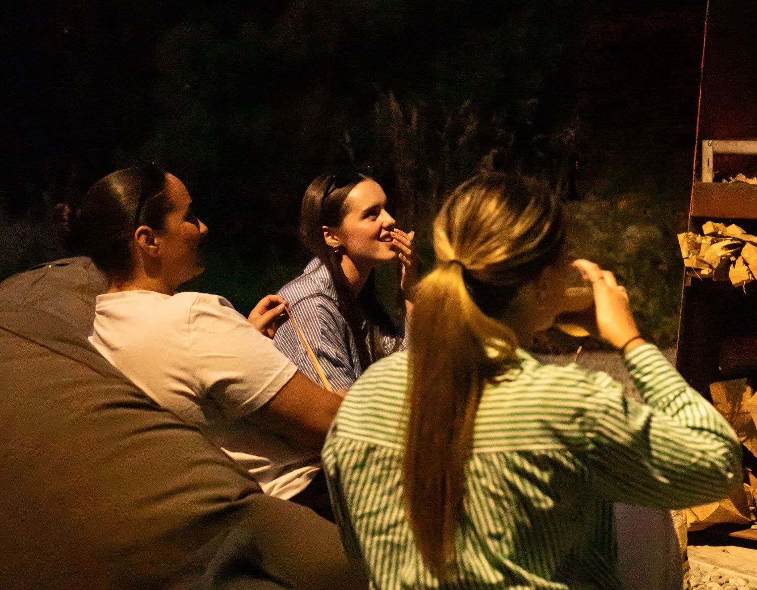 Four women sitting together outdoors at night, engaged in conversation, with a dark background and warm lighting.