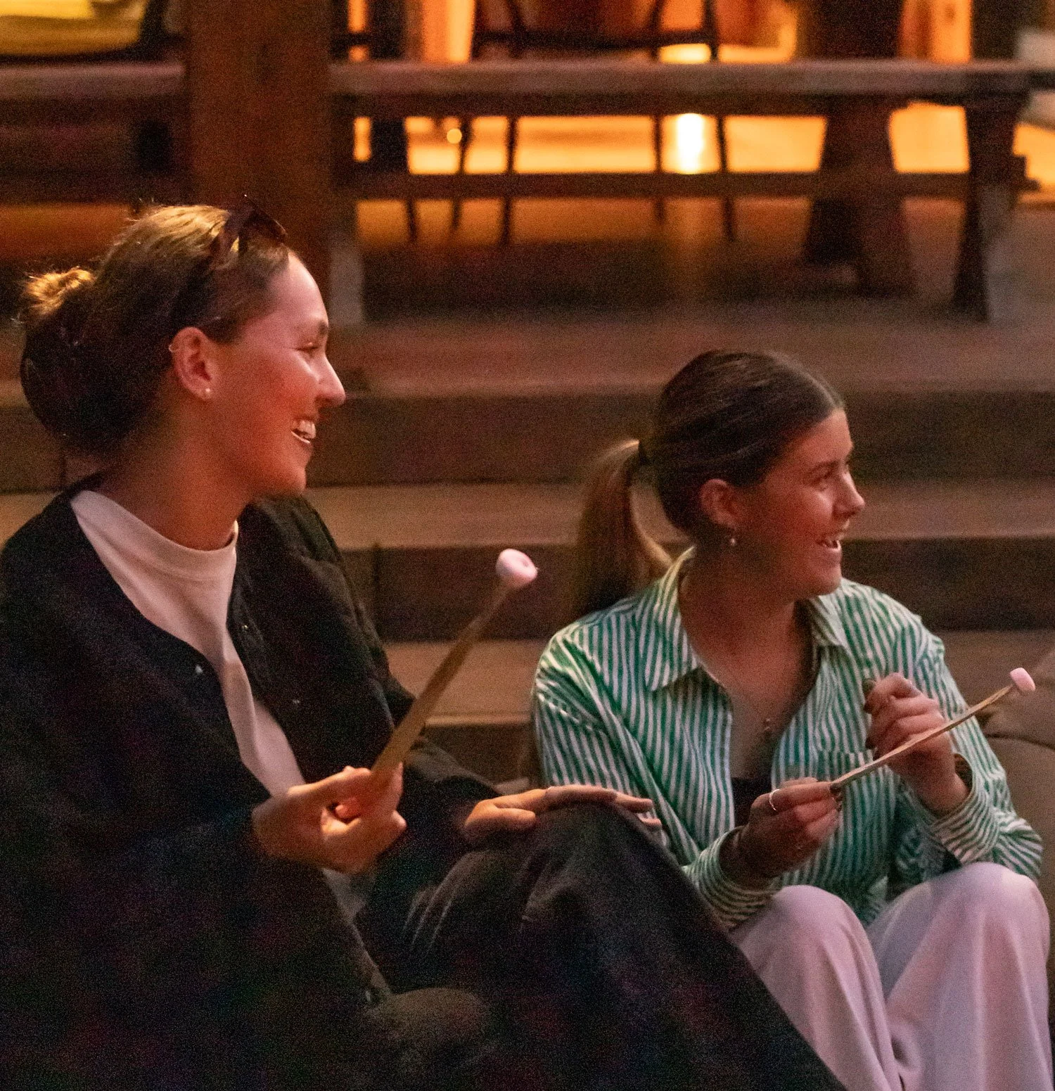 Two women sitting in a cozy, dimly lit cafe, smiling and laughing while holding marshmallow-topped skewers.