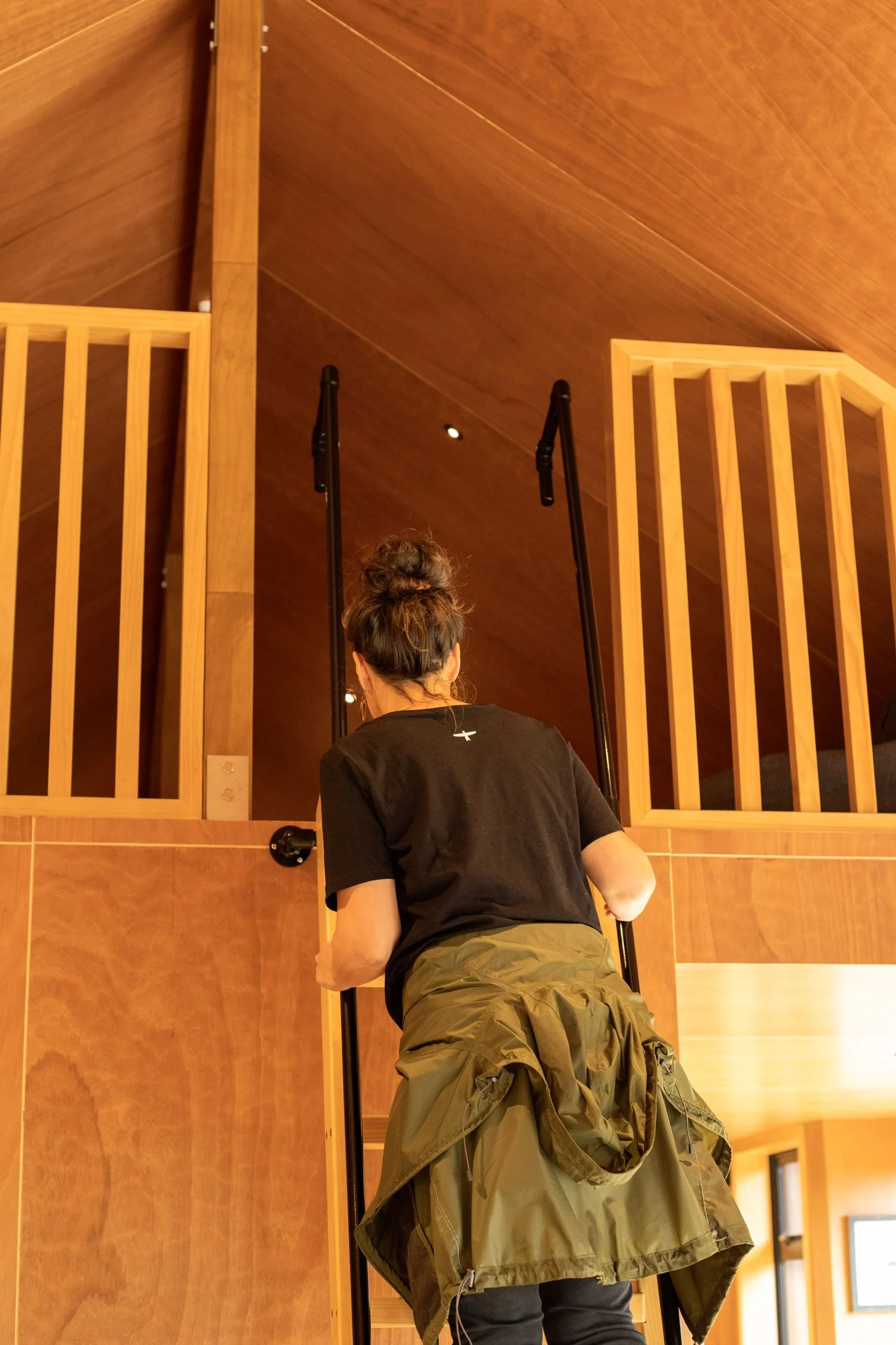 A person with a bun hairstyle, wearing a black t-shirt and khaki pants, is ascending stairs with black handrails. The setting has wooden paneling and railings on either side, with warm lighting from above.