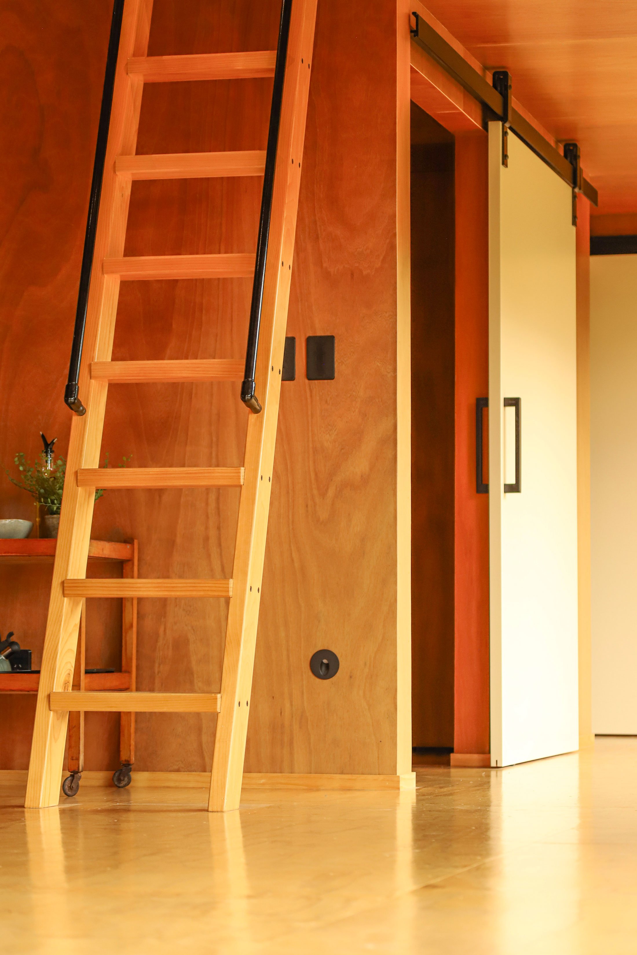 Photo of a wooden kitchen counter with a wooden snack table on wheels and black handles against a wooden wall. The floor is wooden, and there is a white kitchen cabinet with black handles, a sink with a faucet, a plant, and some small items on the co