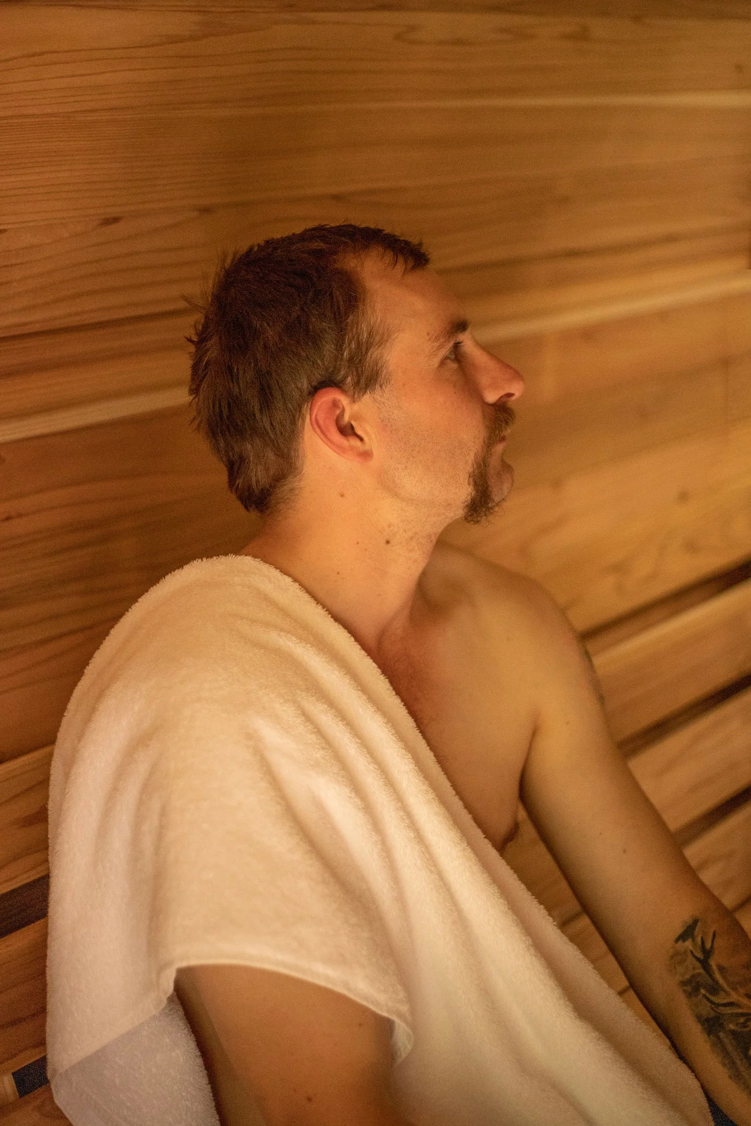 A man with short, brown hair and a goatee, in profile, lying down in a wooden sauna or room, wearing a white towel over his shoulders, with a tattoo of a bird on his upper arm.