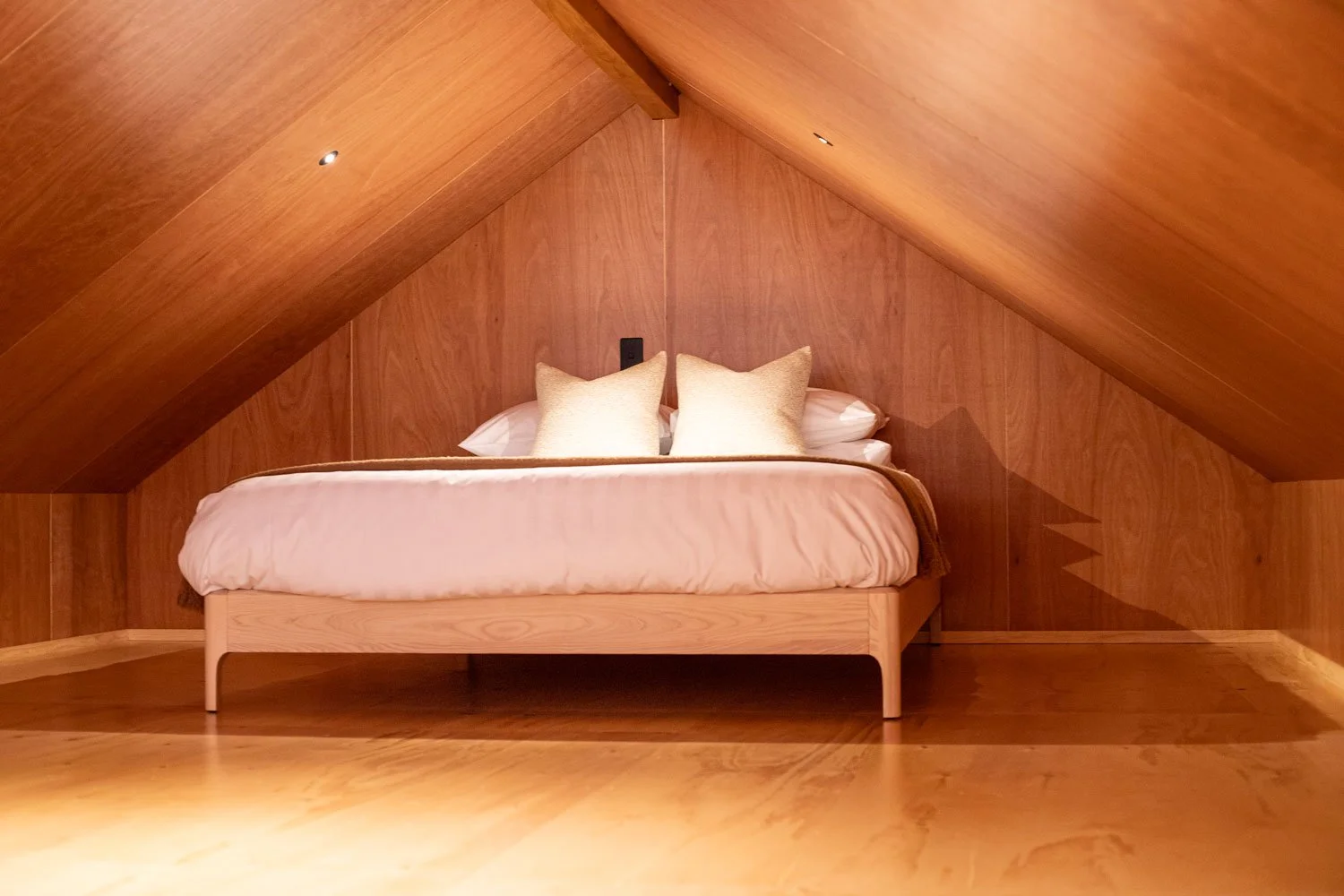 A cozy bedroom in an attic with wooden walls and ceiling, featuring a bed with white bedding and two beige pillows.