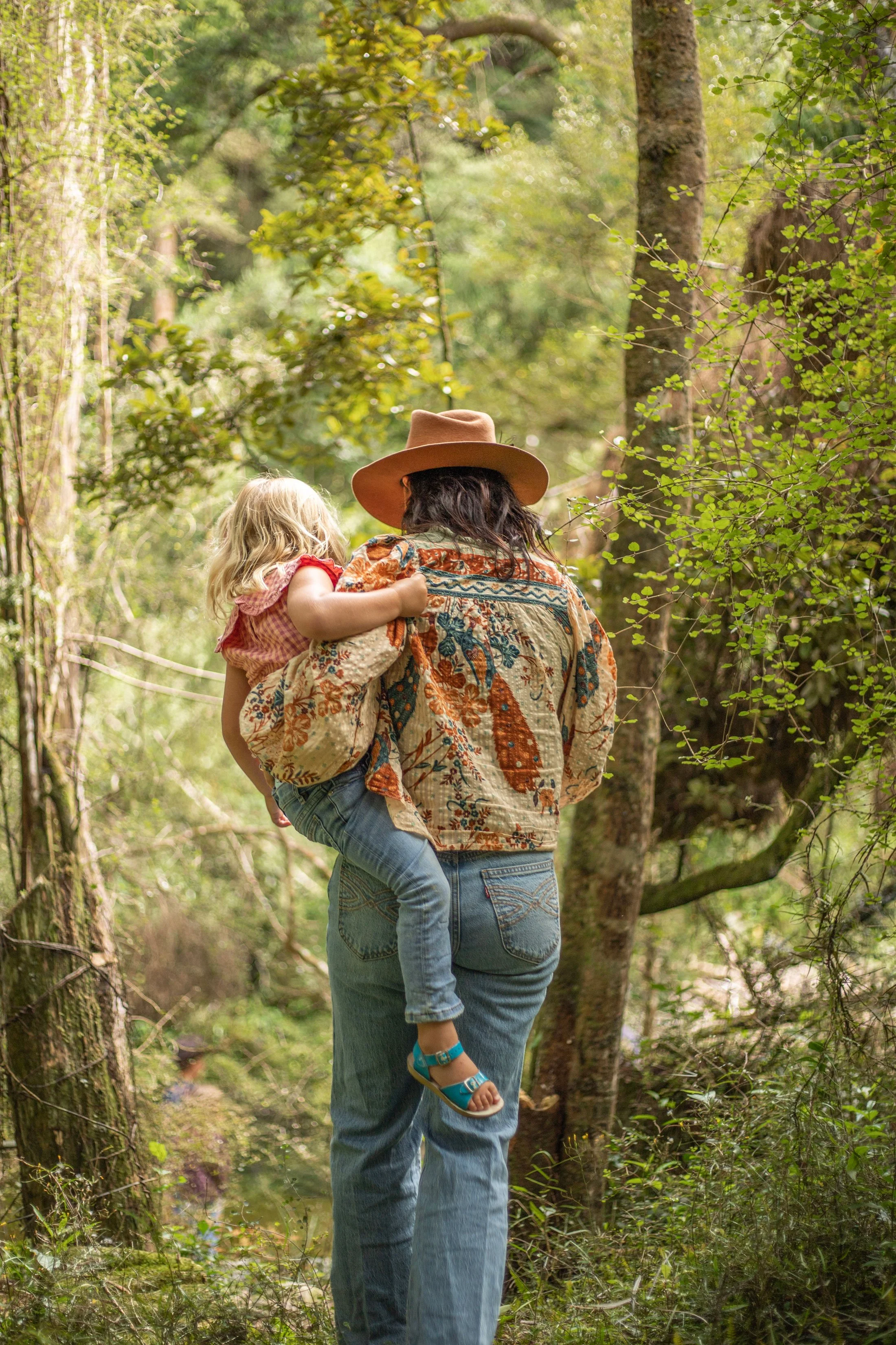 A woman carrying a young girl in a forest with green foliage, trees, and sunlight filtering through the leaves.