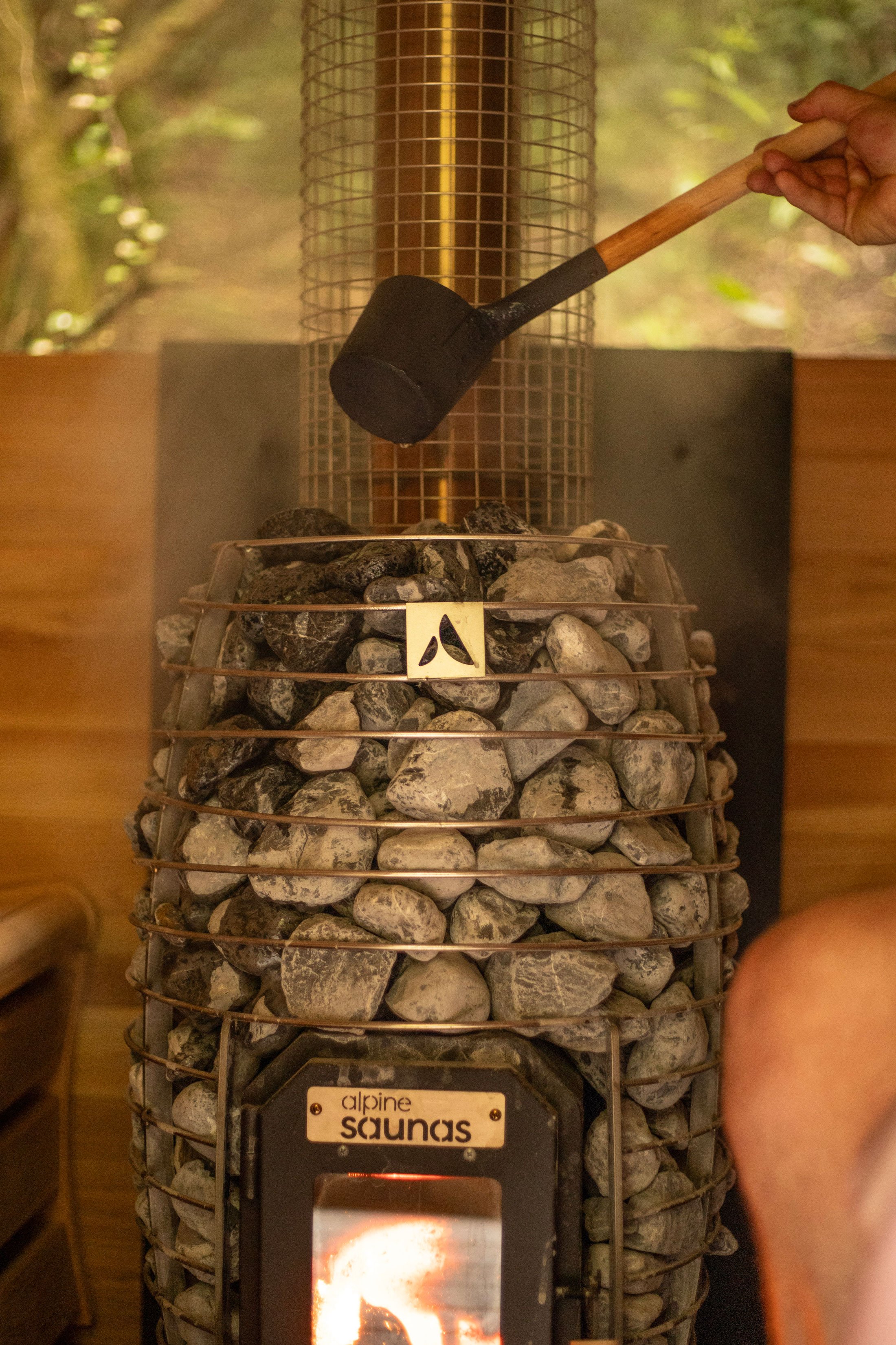 A person using a rubber mallet to strike a pile of gray rocks inside a metal cage, with a heated element at the bottom, in a sauna or similar setting. The background shows a nature scene of green trees.