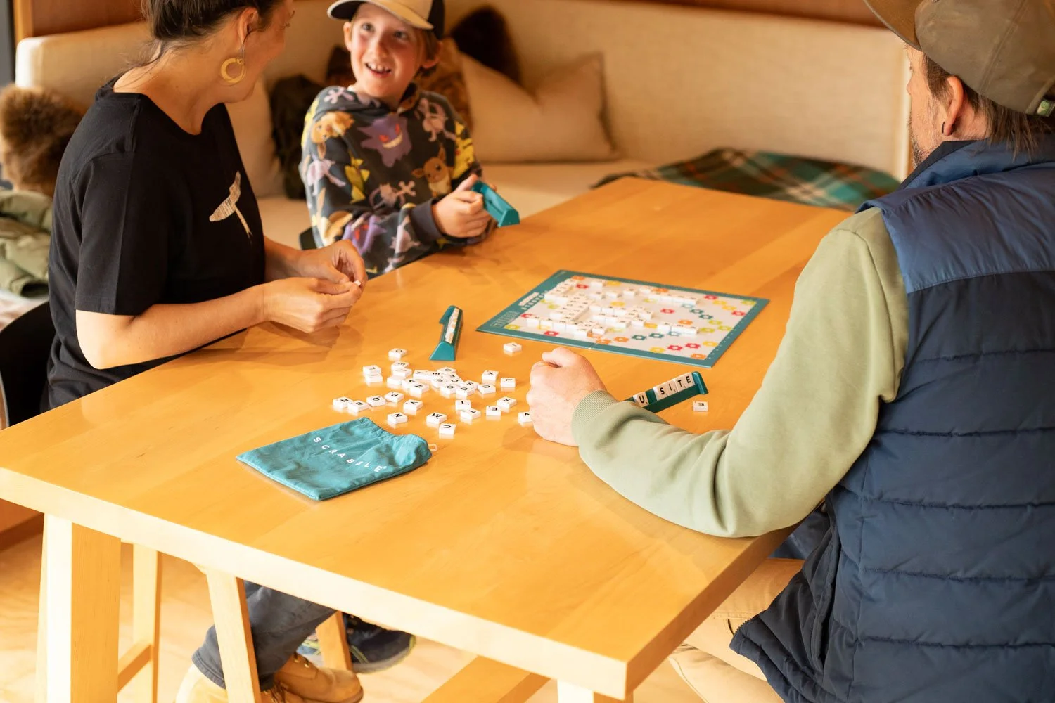 Three people playing Scrabble at a wooden table, with game tiles and a game board visible.