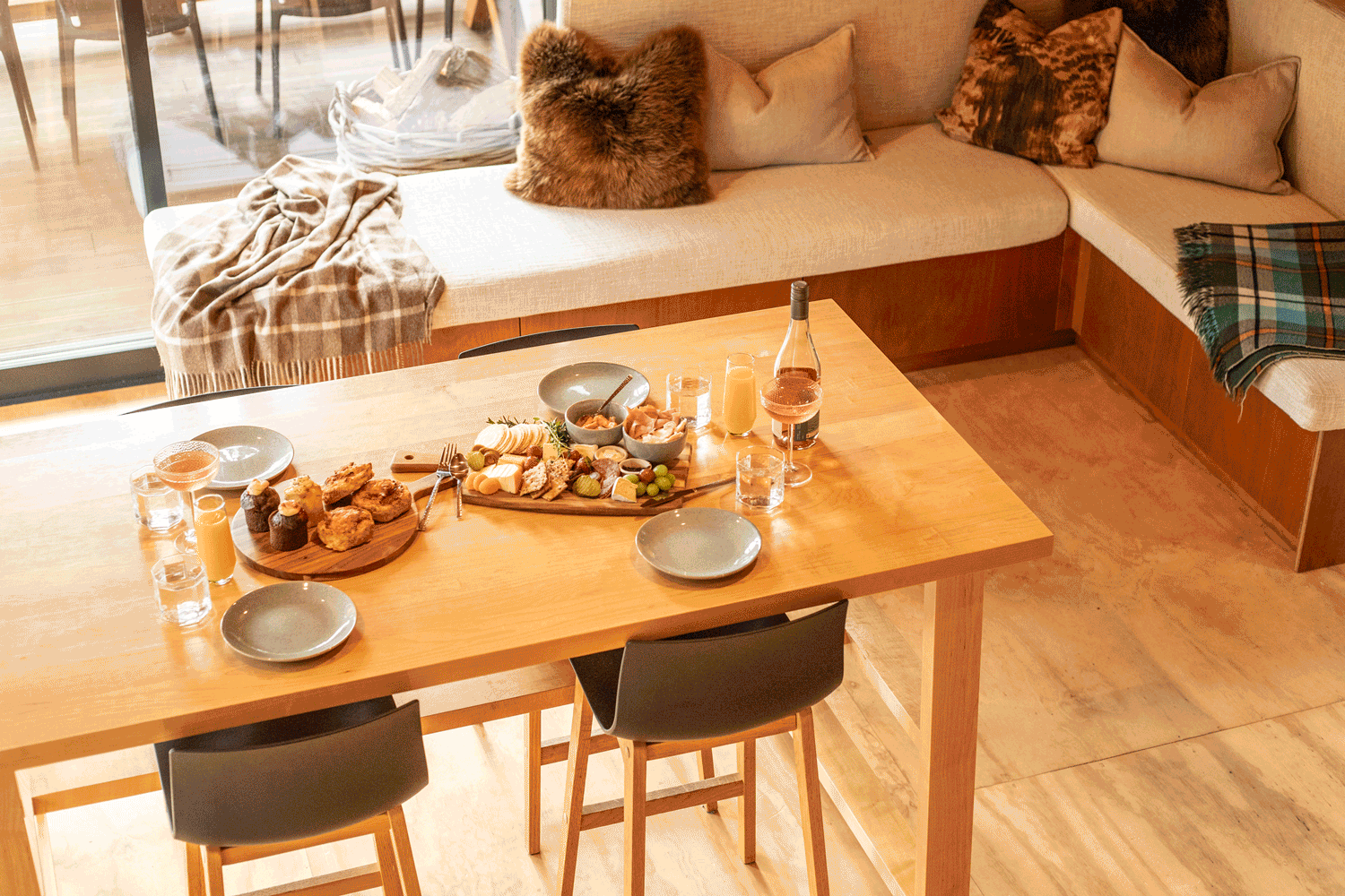 A cozy breakfast scene with a wooden dining table set with various foods including pastries, cheese, grapes, and drinks. In the background, there's a beige sectional sofa with cushions and a plaid throw blanket, situated near a sliding glass door leading to a porch or balcony.
