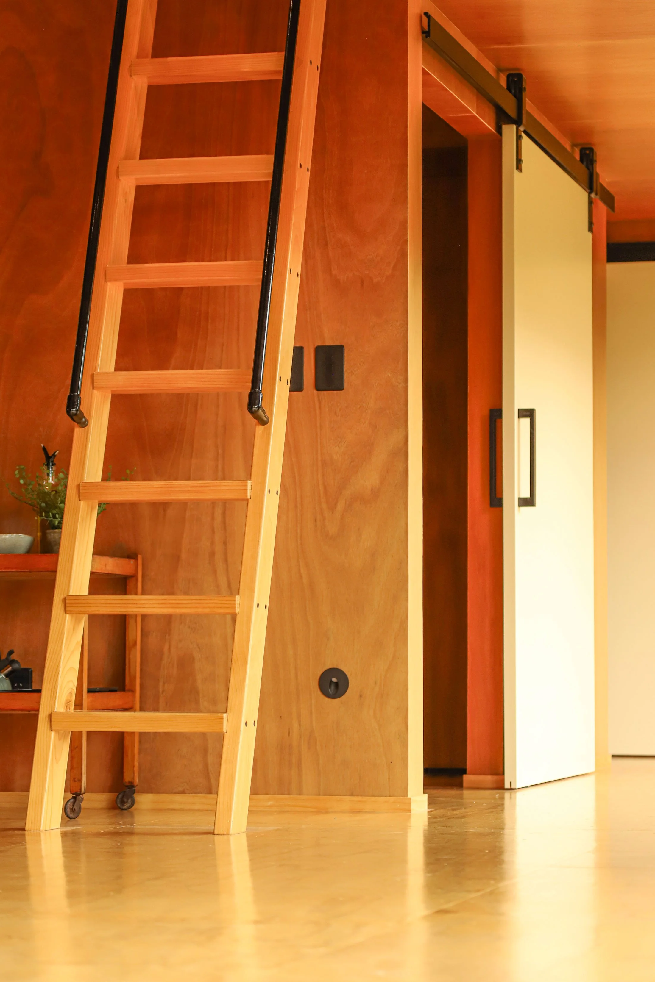 Wooden ladder with wheels leaning against a kitchen island made of wood. The kitchen has white cabinets and a countertop with a potted plant and kitchen faucet. The floor is wood.