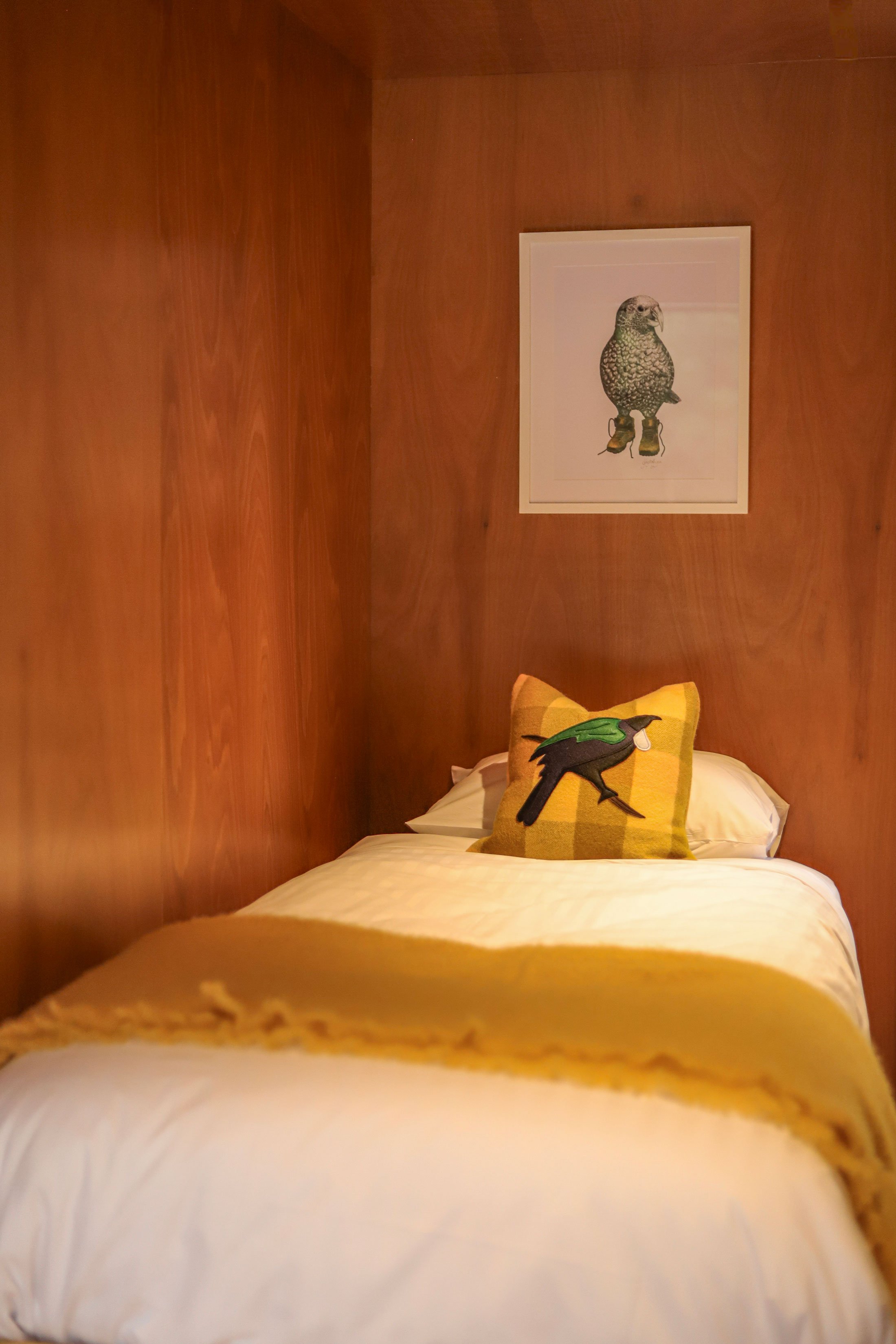 A bedroom with wood-paneled walls featuring a framed picture of an bird, and a bed with a yellow pillow decorated with a bird illustration, and a yellow blanket at the foot of the bed.