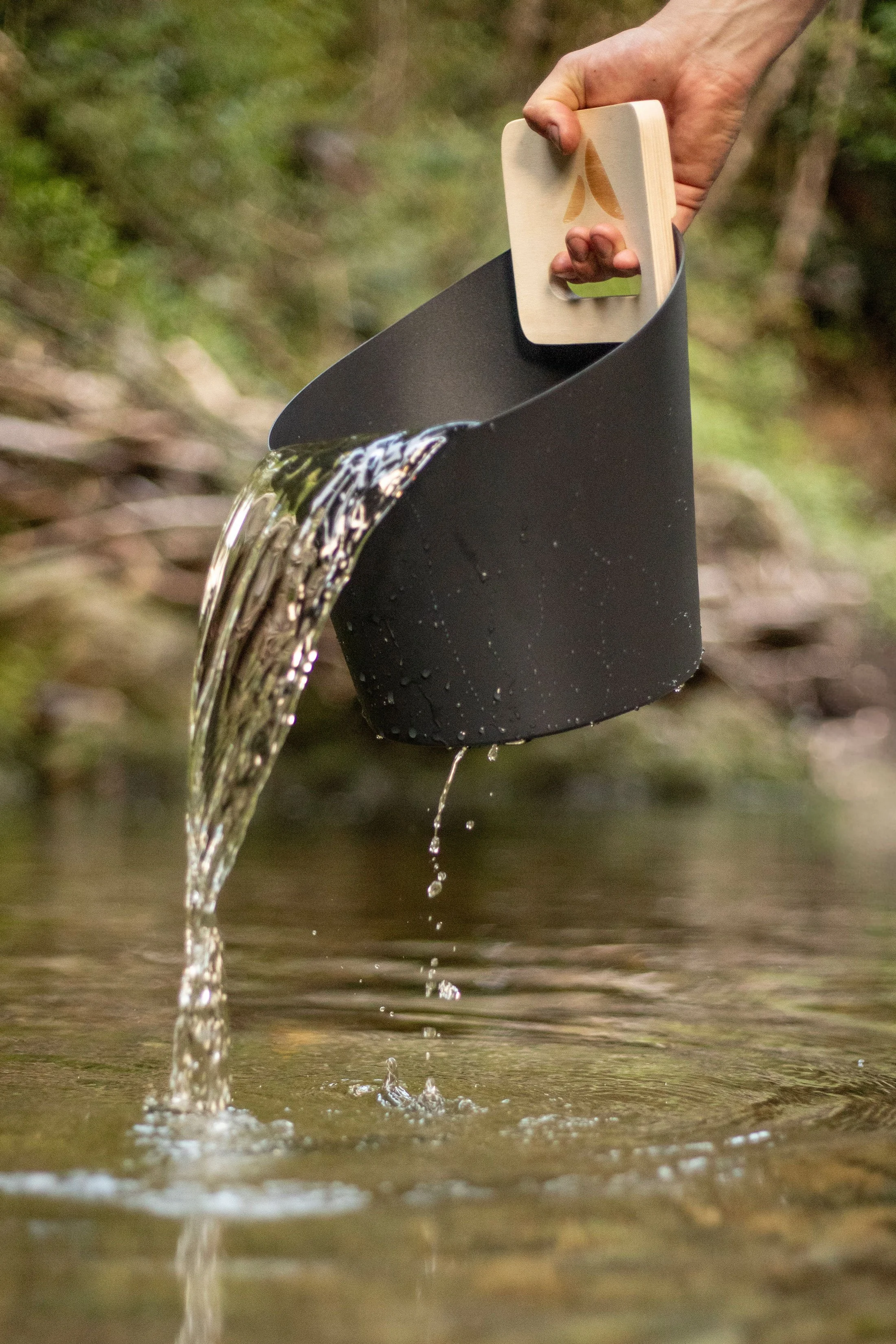 A hand pouring water from a black container with a wooden handle into a body of water.
