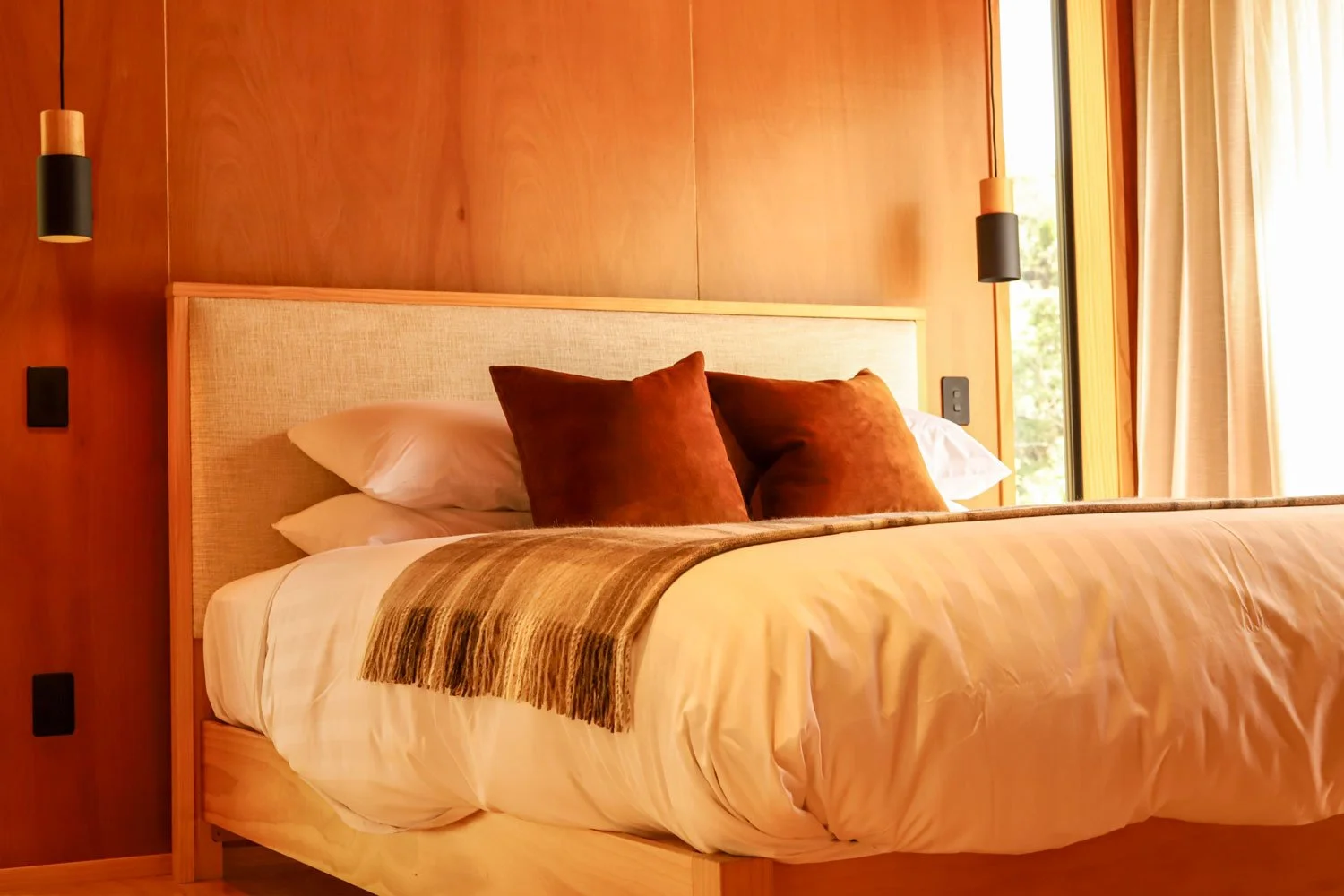 A neatly made bed with two white pillows and three brown pillows, a beige bedspread with a dark brown striped blanket at the foot, in a room with wooden walls, pendant lights, and a large window with beige curtains.
