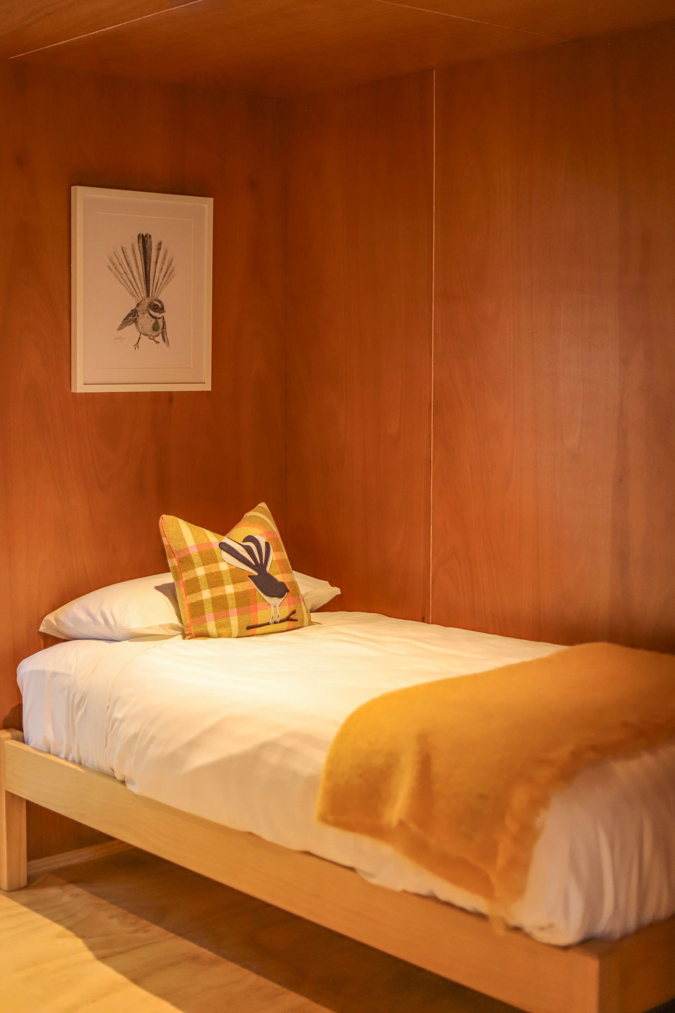 A cozy bedroom with wood-paneled walls, a bed with white linens, a plaid pillow featuring a bird design, and a yellow blanket at the foot of the bed. There is a framed bird illustration on the wall above the bed.