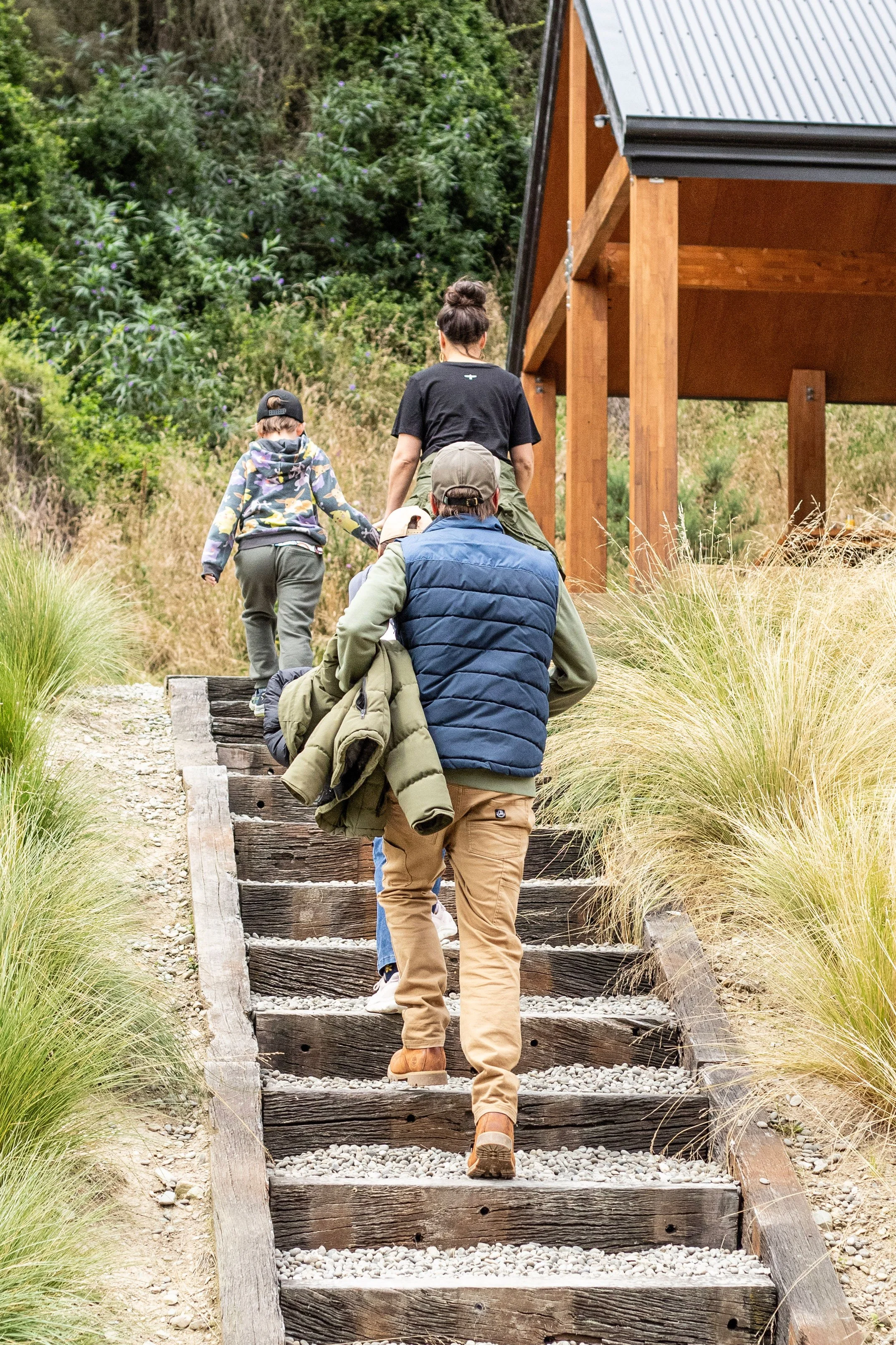 A group of people hiking up outdoor wooden stairs through grassy and bushy landscape with a wooden building in the background.