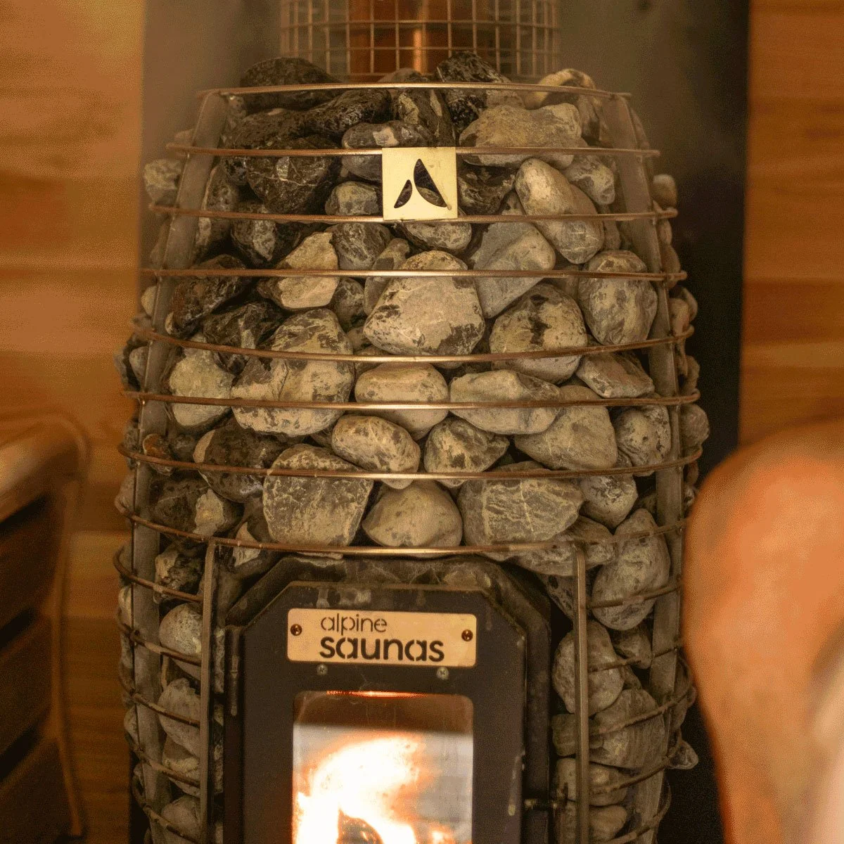 A stone-covered sauna heater with a metal grill on top. The heater has a label reading 'alpine saunas' and a small viewing window showing a fiery interior.
