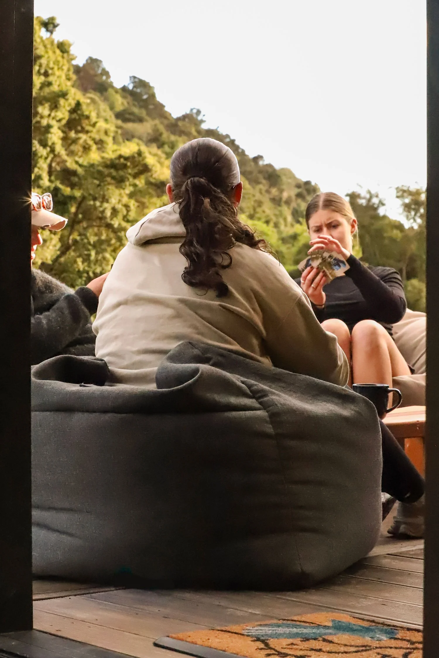 People sitting outdoors around a table with a mountainous, wooded landscape in the background. One person is holding a deck of cards, and another wears glasses with a dog-shaped frame.
