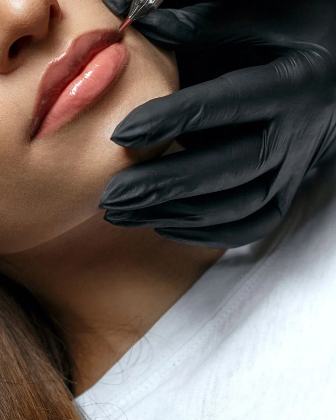 A person receiving a lip tattoo; cosmetic tattooing procedure, with a practitioner wearing black gloves applying pigment to the lips using a fine tool.