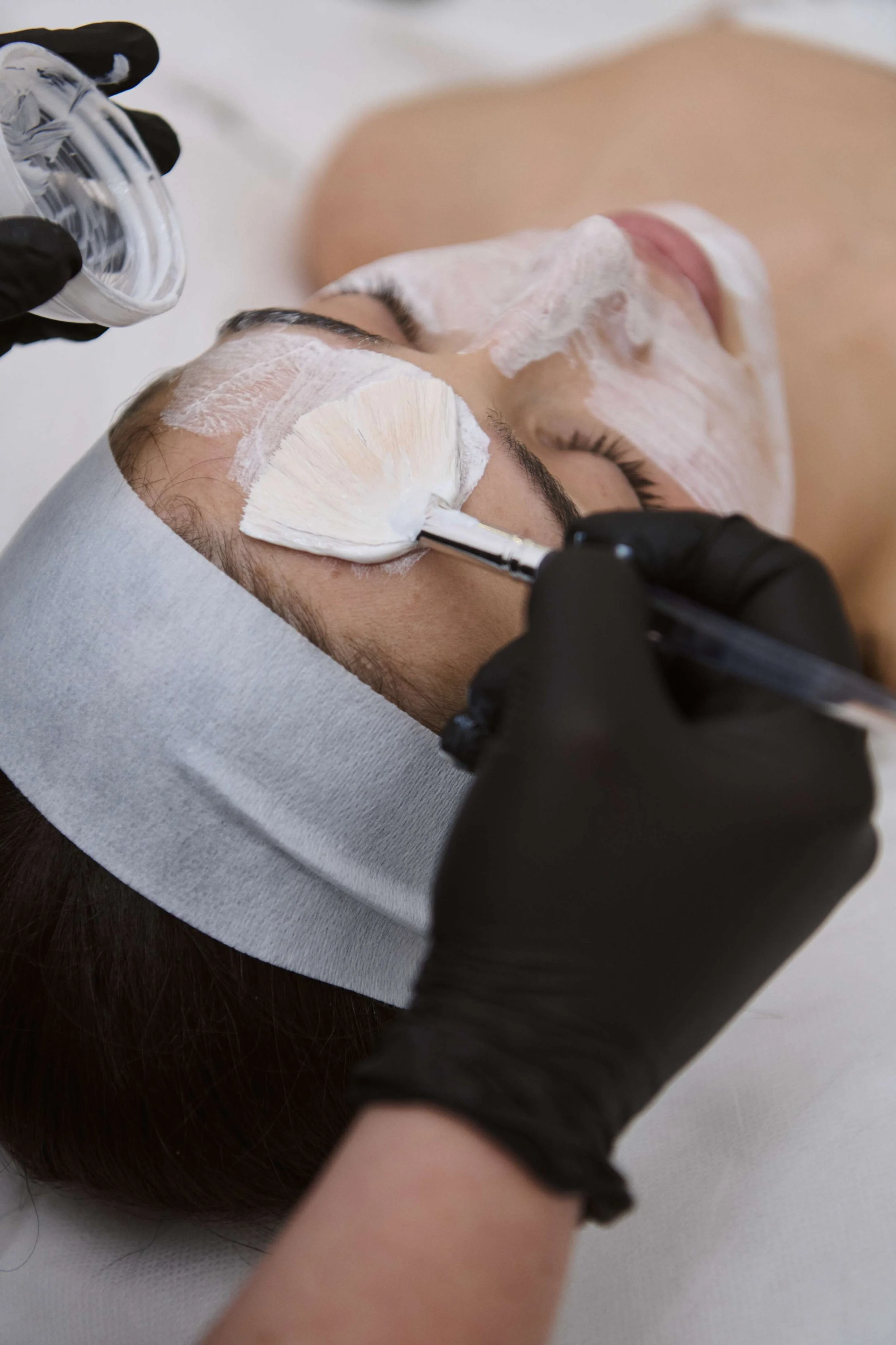 Person lying down receiving a facial treatment, with a facial mask applied and a spatula used to remove or apply a cream, in a spa or skincare setting.