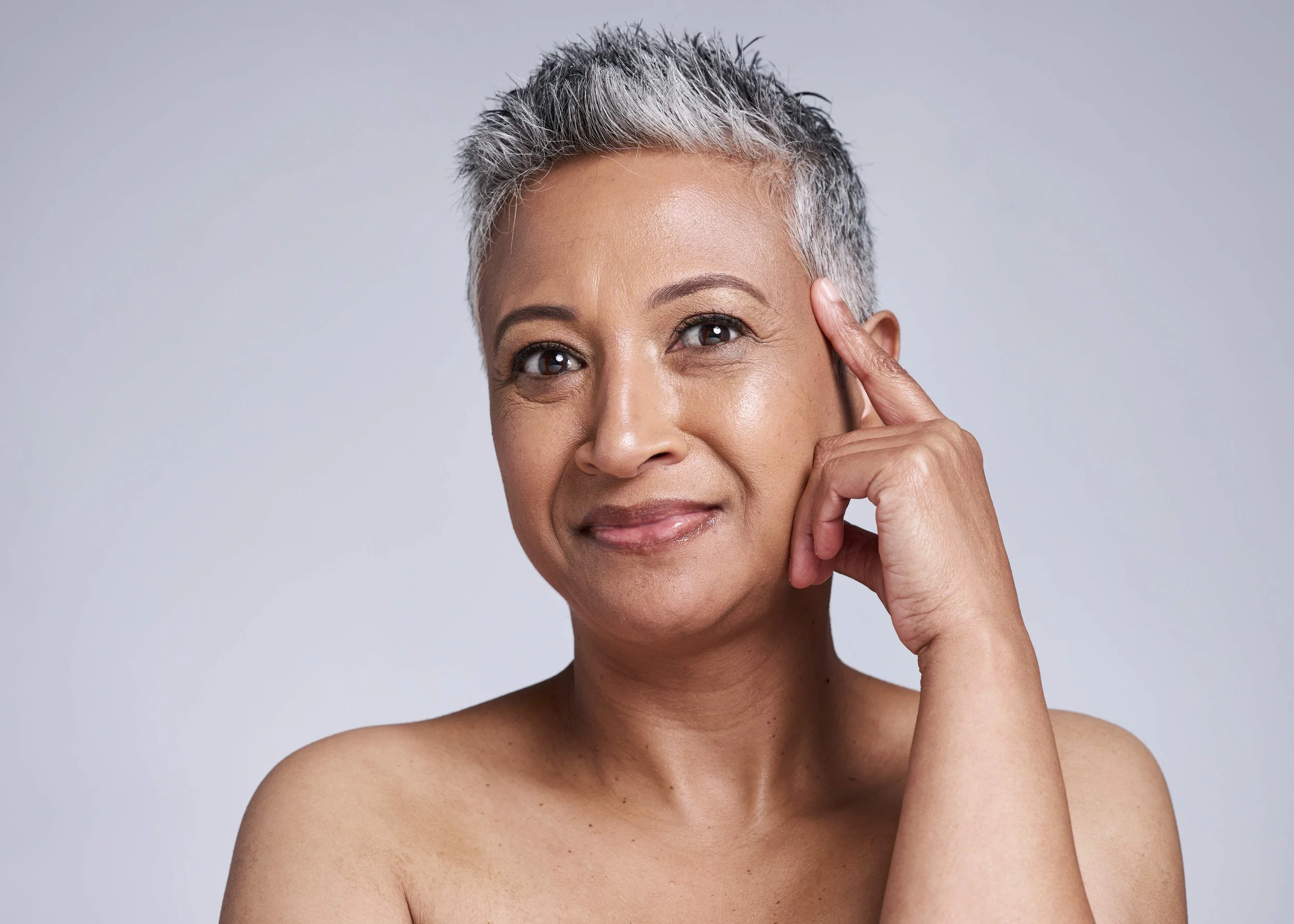 A middle-aged woman with short, spiky gray hair, smiling softly, touching her temple with her index finger, against a plain light gray background.