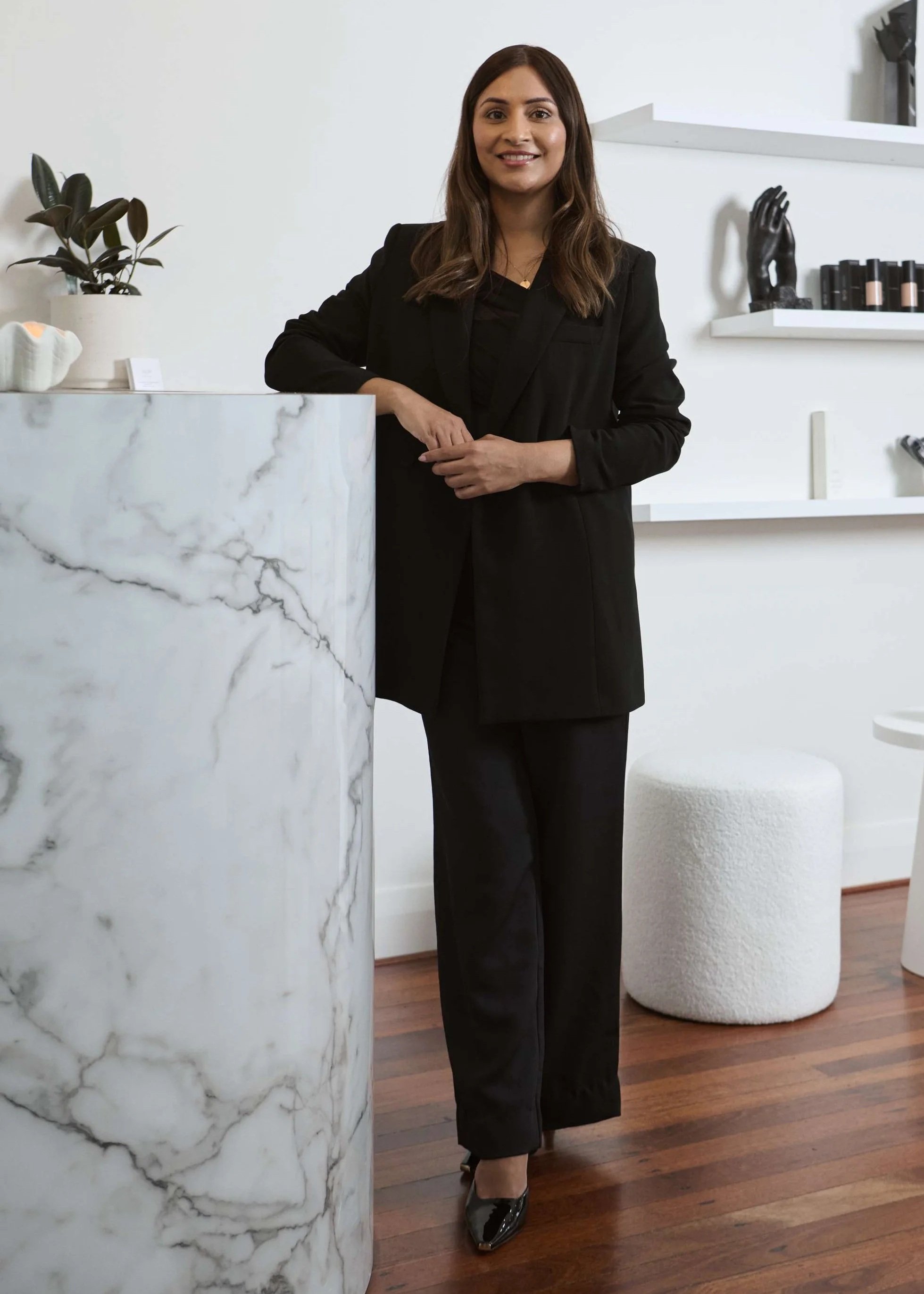 Shelley Reid, Skin Room Perth, wearing a black blazer, black top, and black wide-leg pants, standing in a modern, minimalist interior near a marble countertop with a potted plant, with a white wall with shelves and decorative items in the background.