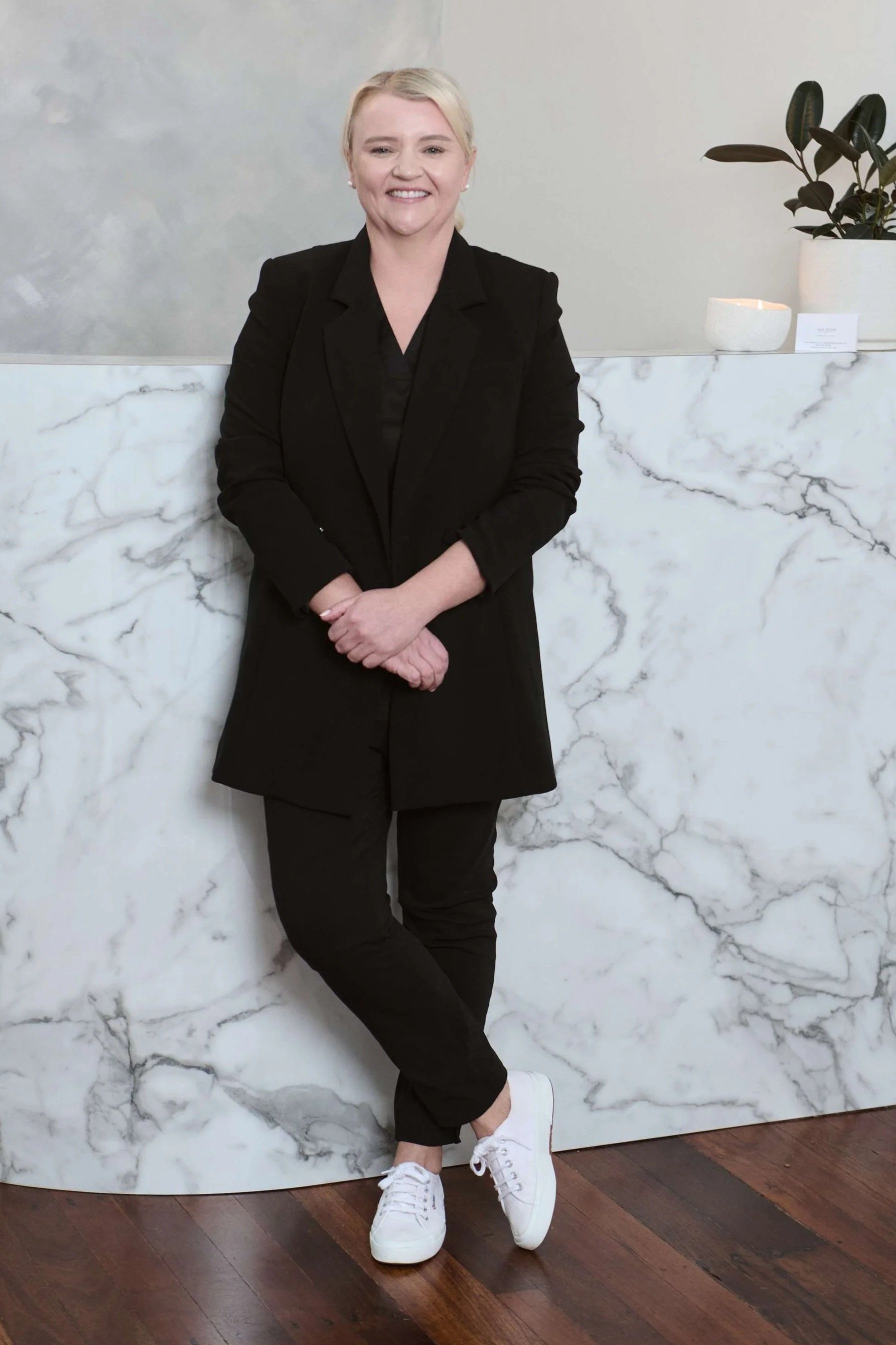 Amy Middleton | Senior Dermal Therapist and Women's Health and Wellness Coach, Skin Room Perth, dressed in a black blazer and pants, wearing white sneakers, stands against a marble wall with wooden flooring.
