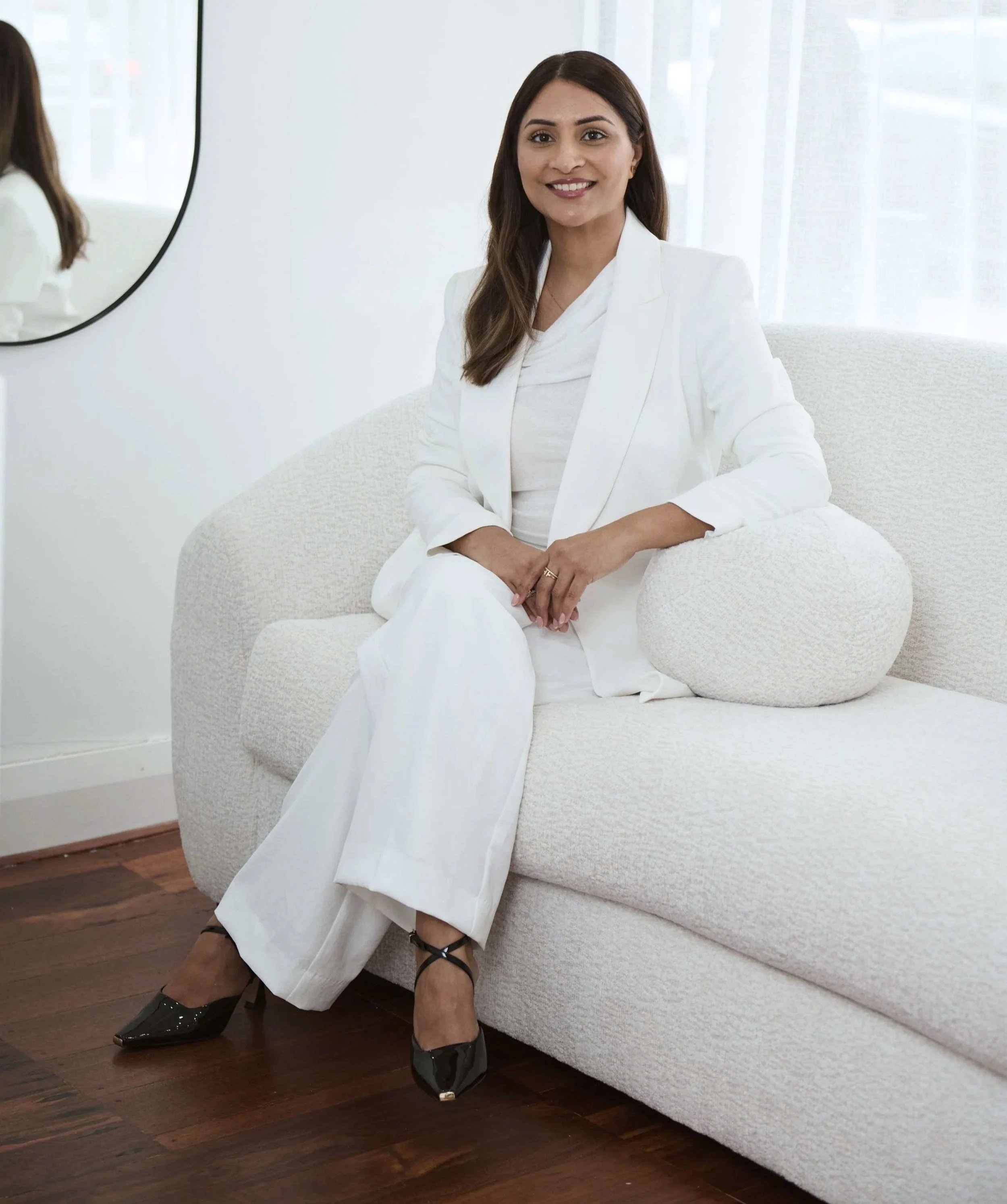 Shelley Reid of Skin Room Perth, sitting on a white textured sofa near a wall with windows, smiling, wearing a white blazer, white pants, and black high-heeled shoes, with her hands clasped on her lap.