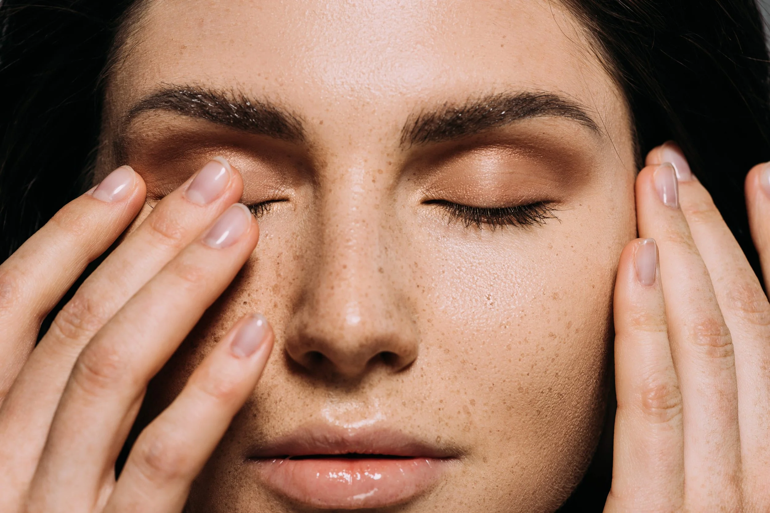 A close-up of a woman with dark hair, closed eyes, and fingers touching her eyelids, possibly applying or removing makeup.