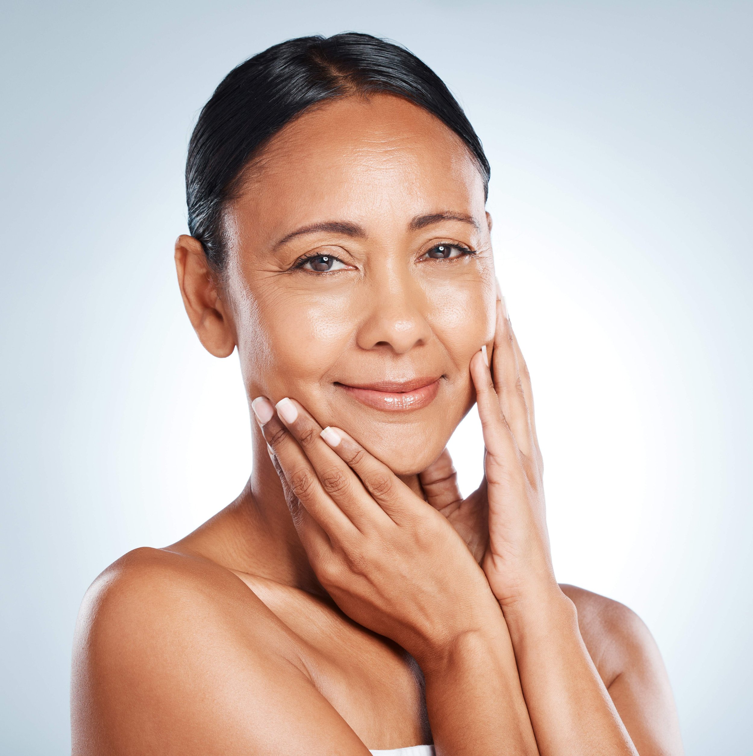 Close-up of a smiling woman with short, dark, wet hair and tan skin, touching her face with both hands, against a light gray background.