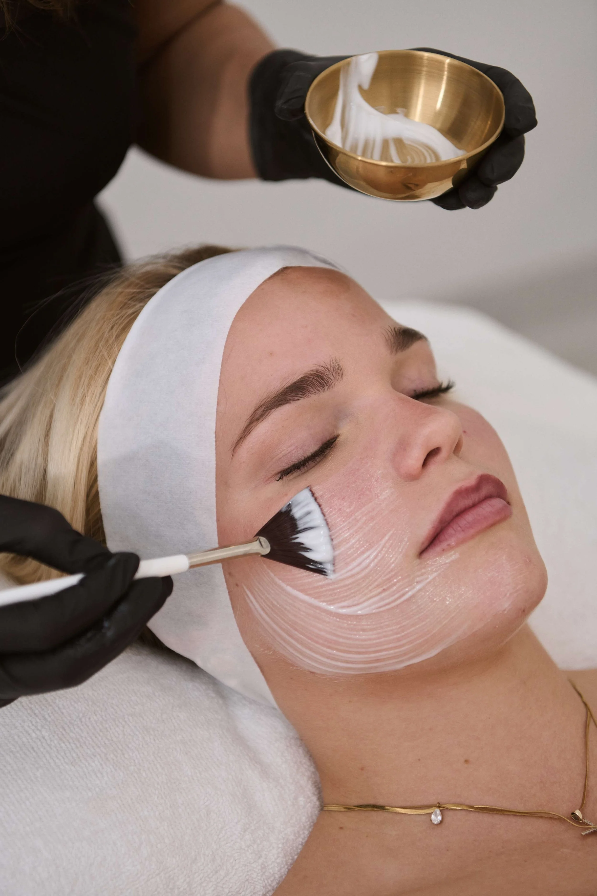 A woman with a white headband is lying down with her eyes closed while a skincare professional applies a white cream or mask to her face using a brush. The professional is wearing black gloves and holding a gold bowl filled with the cream.