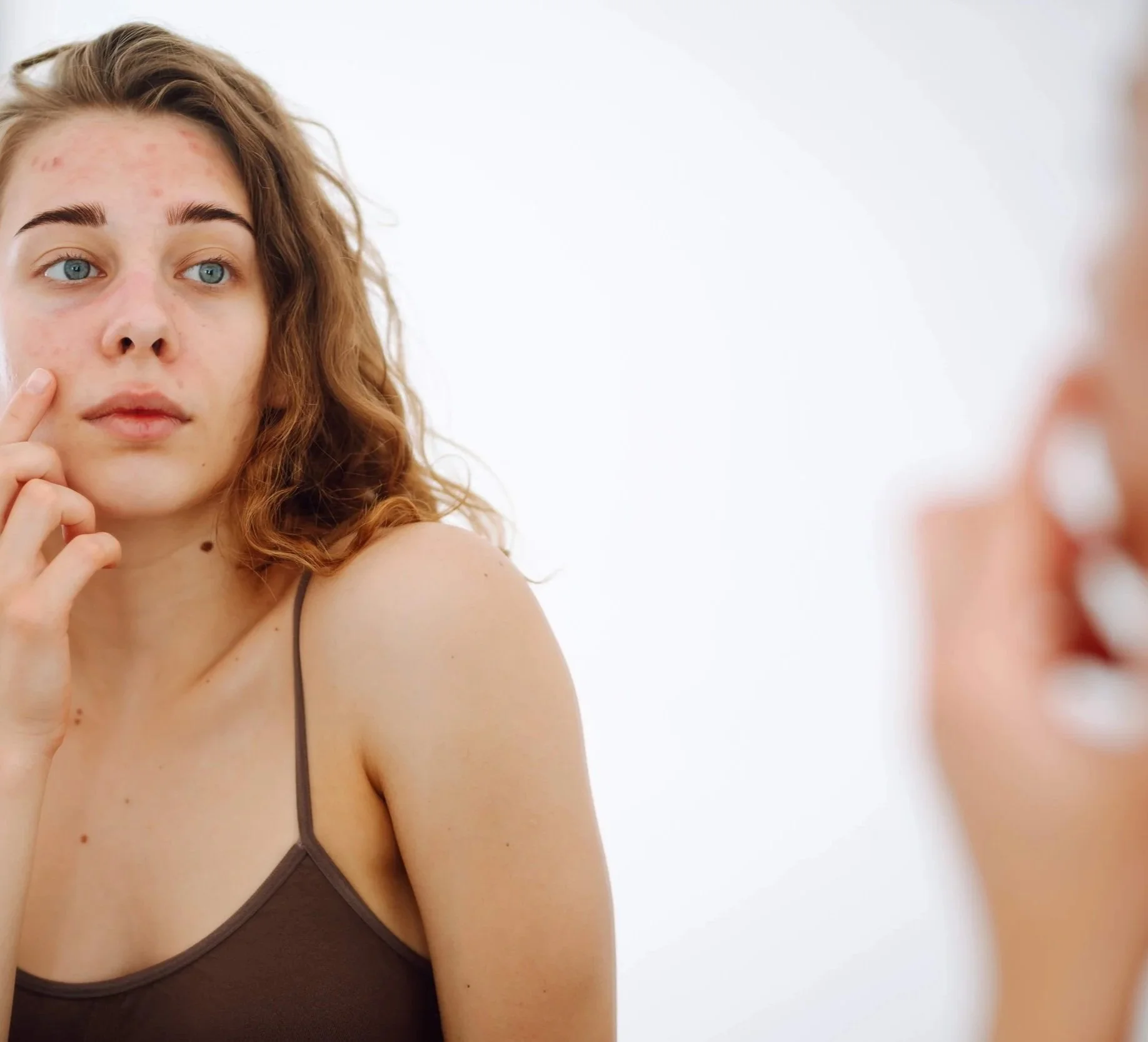 A young woman with curly red hair and blue eyes gazes into a mirror, touching her face. She is wearing a brown spaghetti strap top and is in a well-lit room with a white background.