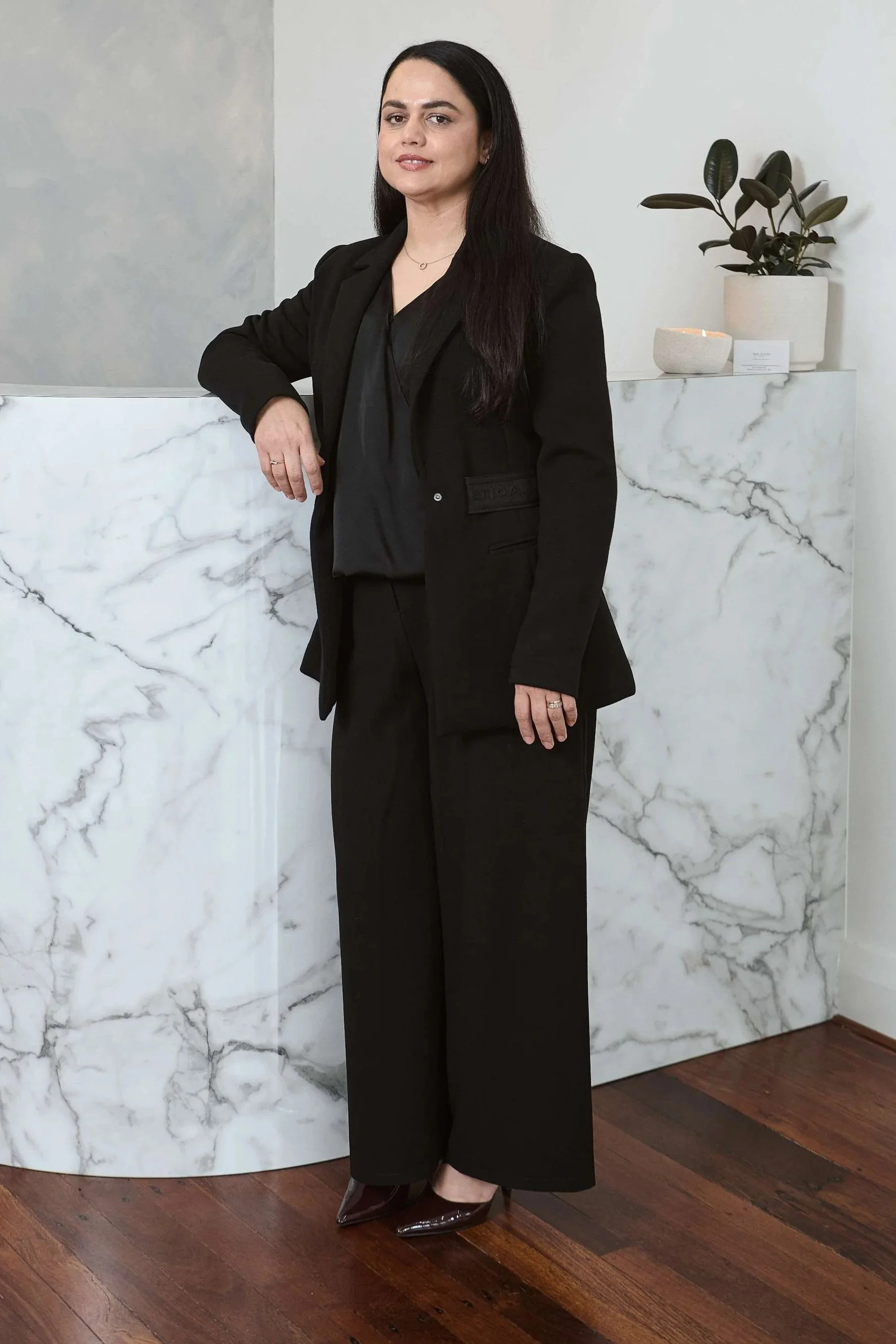 Mushda Musawer | Cosmetic Nurse with over 18 years of experience in the medical industry, Skin Room Perth, with long dark hair dressed in a black blazer, black blouse, and black wide-leg pants standing in front of a marble wall.