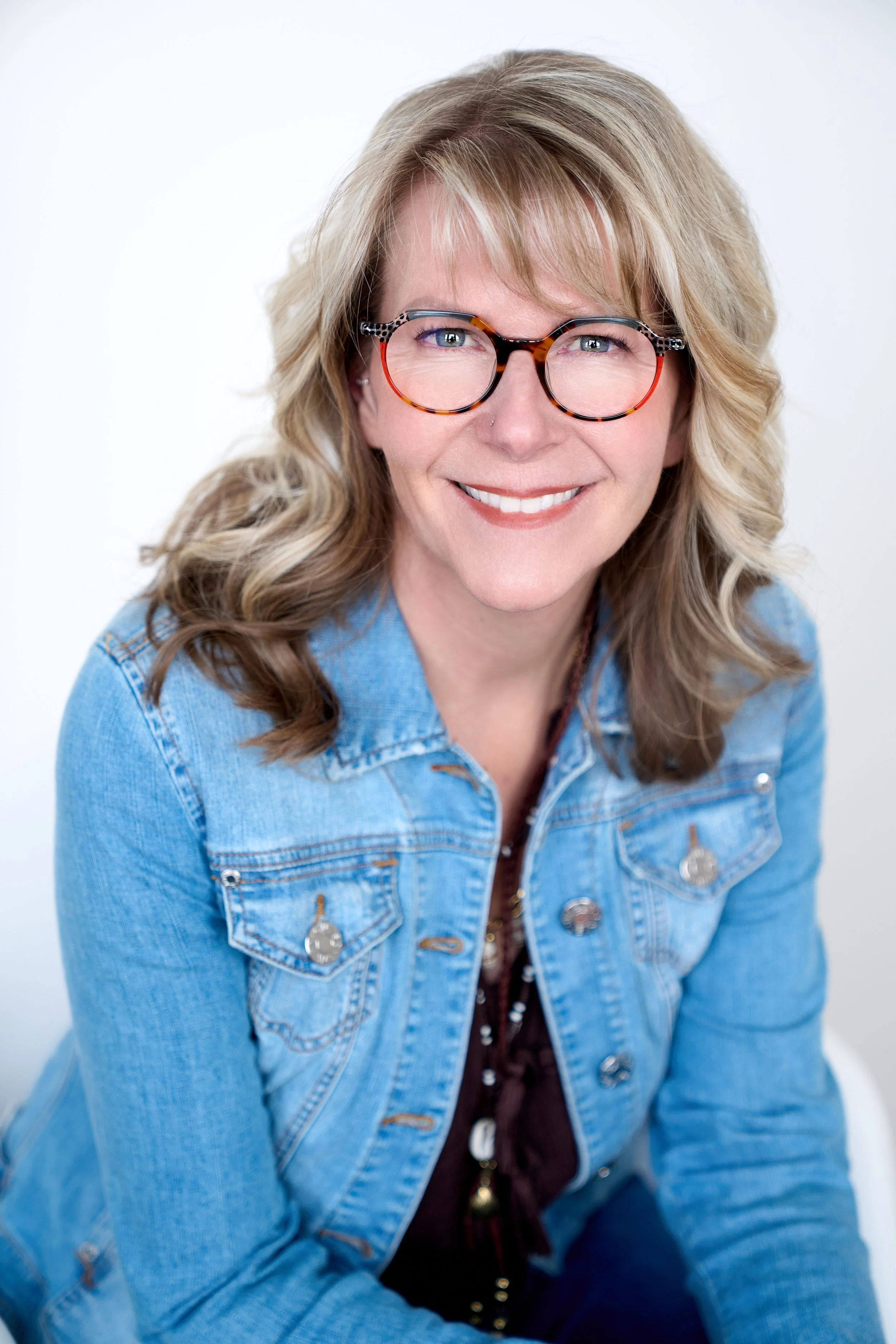 A smiling woman with blond wavy hair wearing glasses, a denim jacket, and a patterned blouse against a white background.