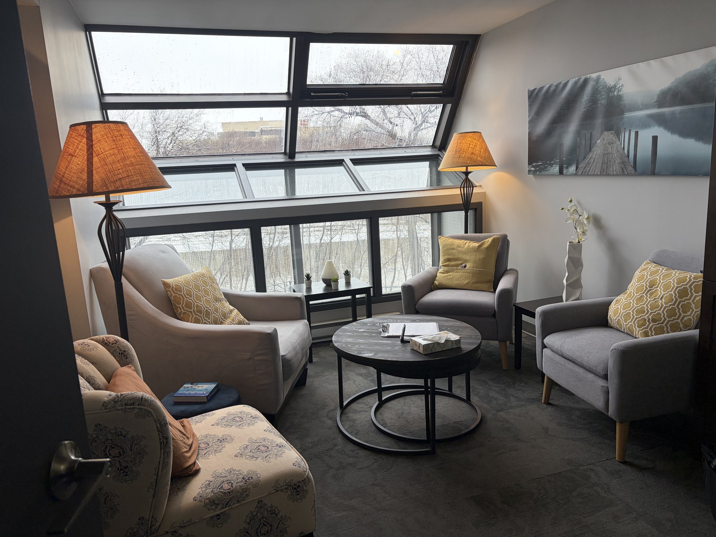 A cozy sitting area with large windows that have rain droplets on the glass, overlooking a leafless tree and distant buildings. The room features two beige armchairs with yellow patterned cushions, a small round coffee table with a notebook and glasses, and a tall white vase with white flowers. Two table lamps with orange shades provide warm lighting. A large landscape photograph of a dock extending over water hangs on the wall.