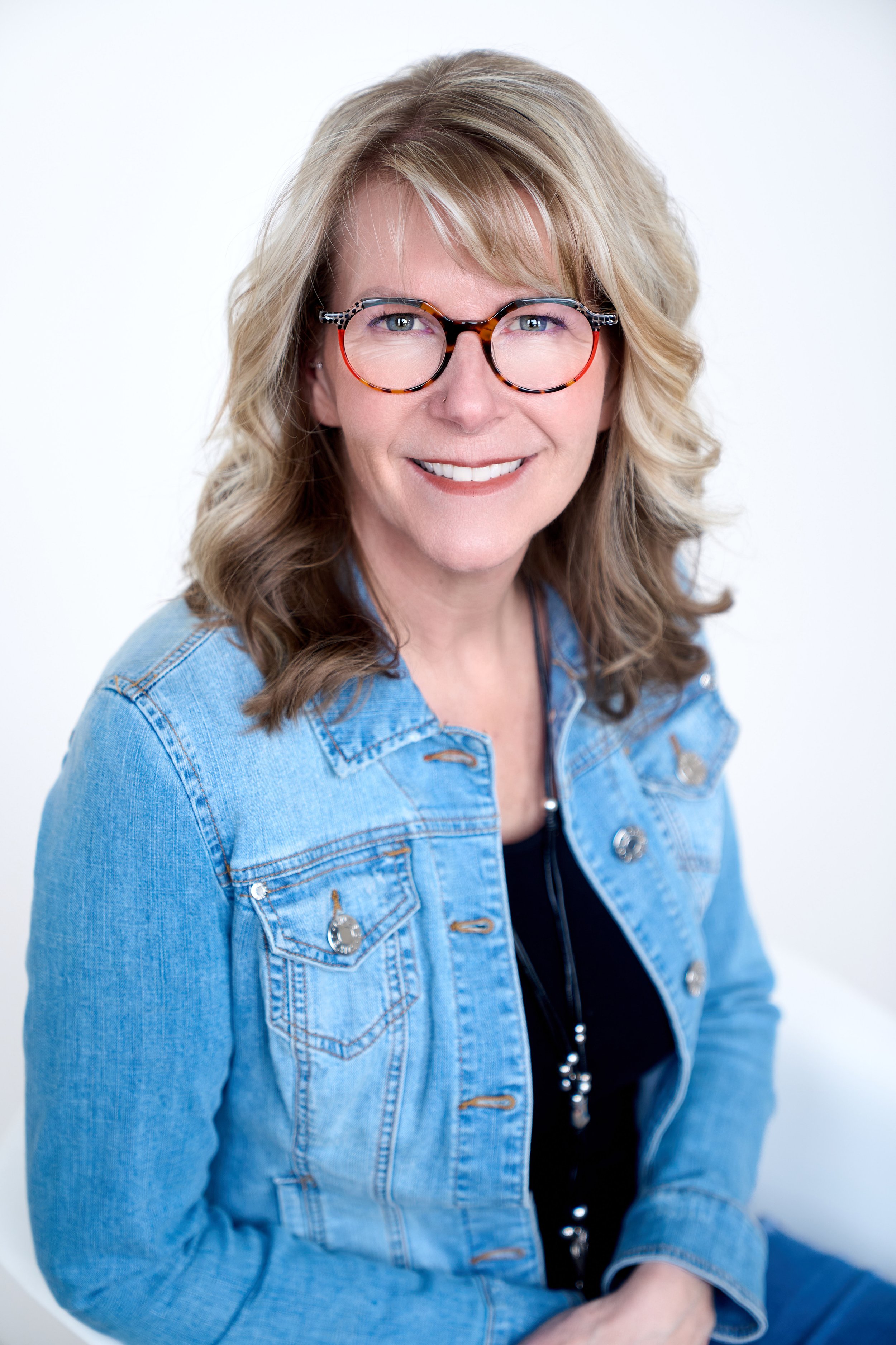A middle-aged woman with blonde, wavy hair and glasses smiling at the camera, wearing a blue denim jacket over a black top, against a plain white background.
