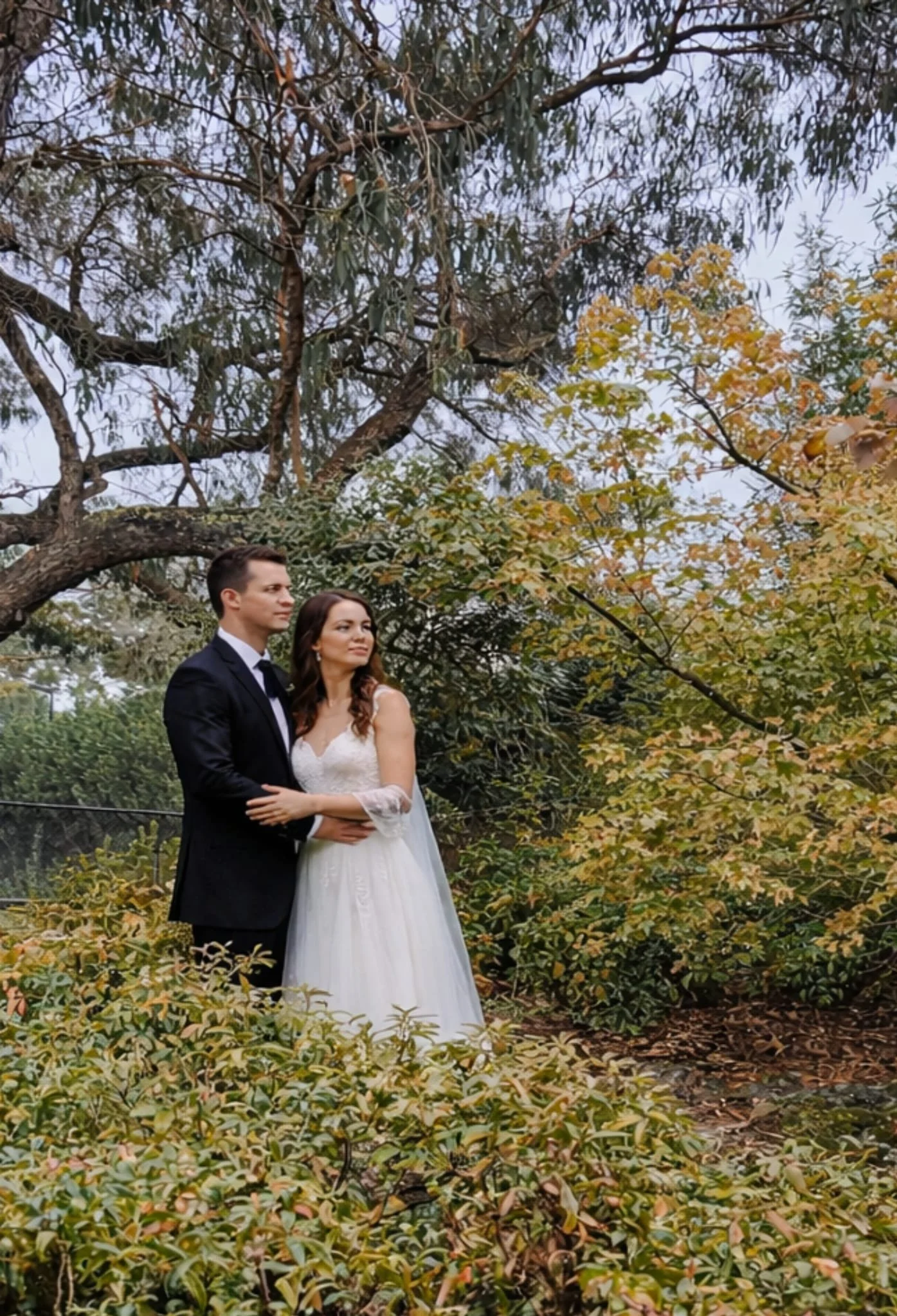 A newlywed couple in wedding attire standing outdoors among trees and bushes with autumn leaves.