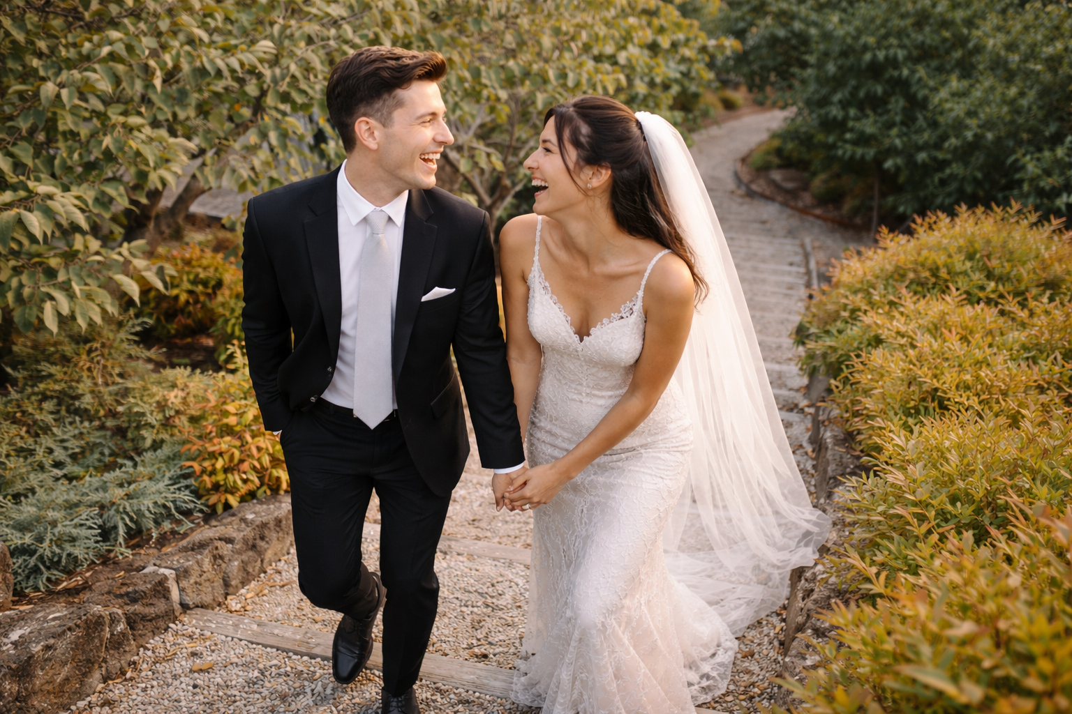 A newlywed couple holding hands and laughing while walking on a garden path surrounded by green and yellow foliage.