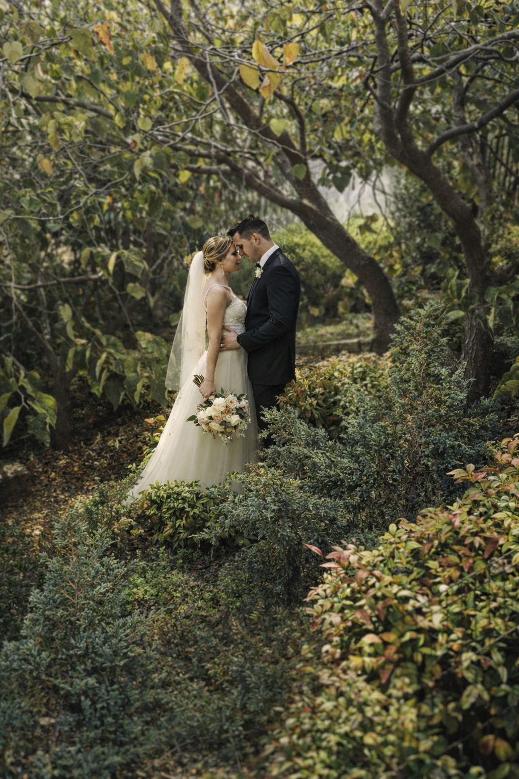 A bride and groom embrace in a lush, green garden, standing close with their foreheads touching. The bride wears a white wedding gown and veil, holding a bouquet of pastel flowers. The groom wears a dark suit. Surrounding them are trees and bushes, c