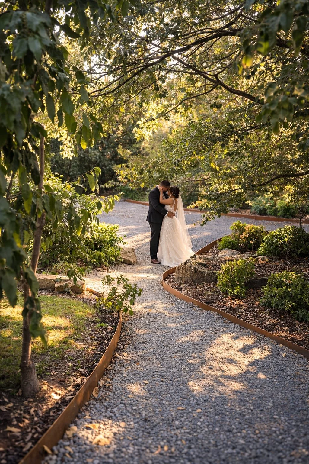 A bride and groom embrace and kiss on a gravel pathway in a lush green garden during sunset.