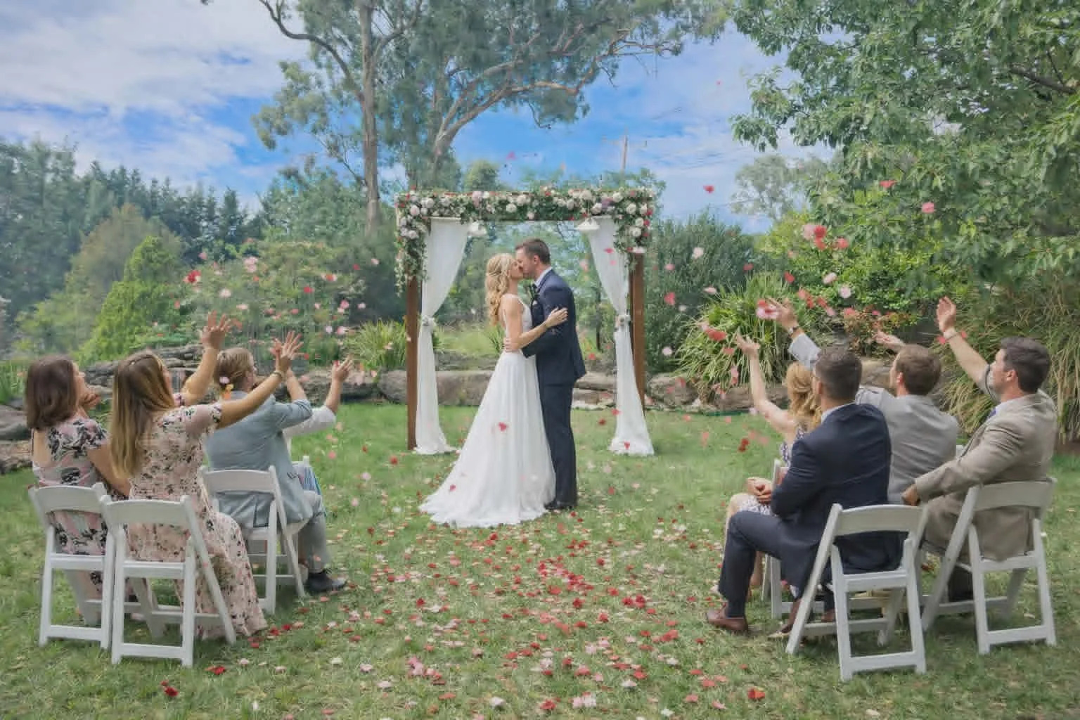 A couple kisses under a wedding arch decorated with flowers during an outdoor wedding ceremony. Six guests seated on either side of the aisle are tossing flower petals in celebration.