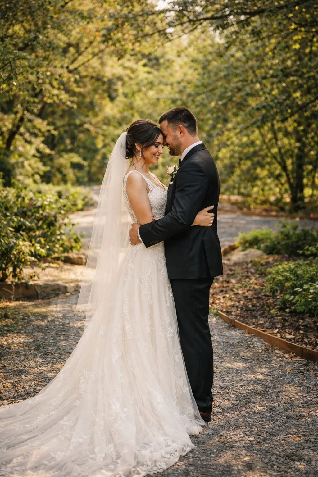A bride and groom in wedding attire standing close, with foreheads touching and eyes closed, in a scenic outdoor setting during daytime.