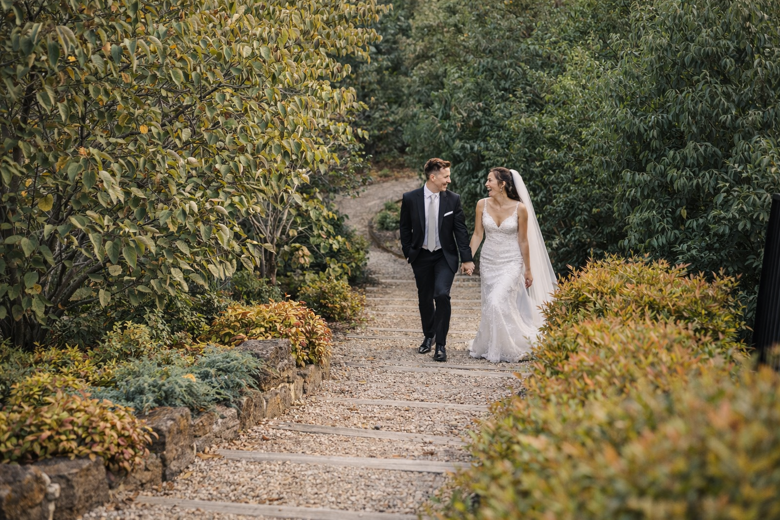 A newlywed couple walking hand in hand on a landscaped garden pathway surrounded by lush green bushes and trees, the bride wearing a white lace wedding gown with a veil and the groom in a black suit with a white shirt and tie, both smiling at each ot